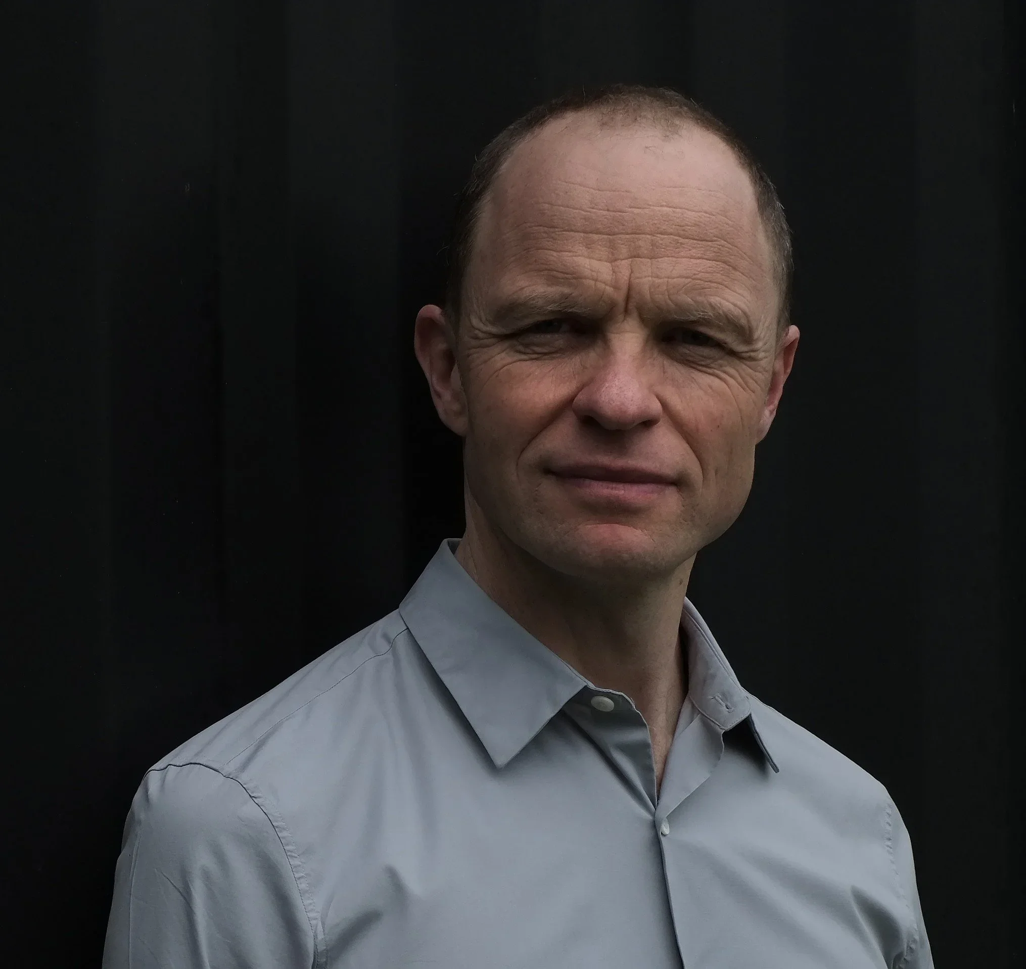 A middle-aged man with short hair and a light gray dress shirt standing against a black background, looking slightly to the side.