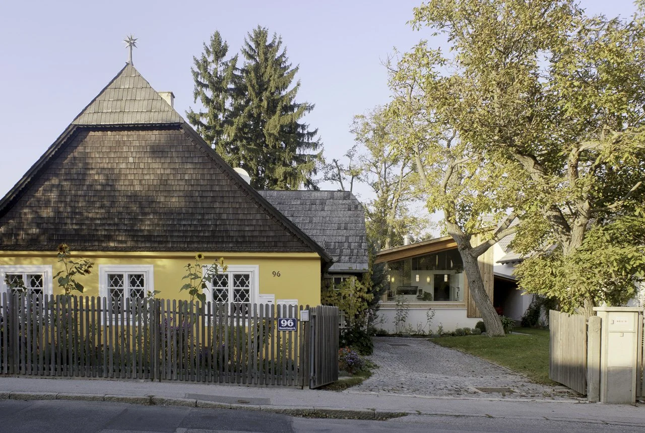 A yellow house with a brown tiled roof, white-framed windows, and a black picket fence, situated on a corner with a driveway, next to a modern white house with large windows and a sloped roof, surrounded by trees.