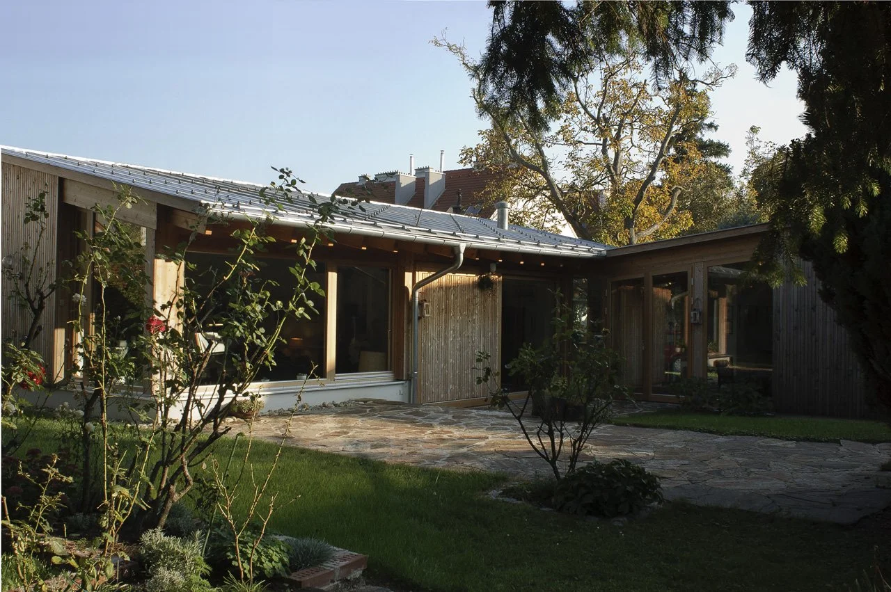 A modern house with large windows, surrounded by a well-kept green lawn, small trees, and a stone pathway. The house has a wooden exterior and a sloped metal roof, with a background of tall trees and a clear blue sky.