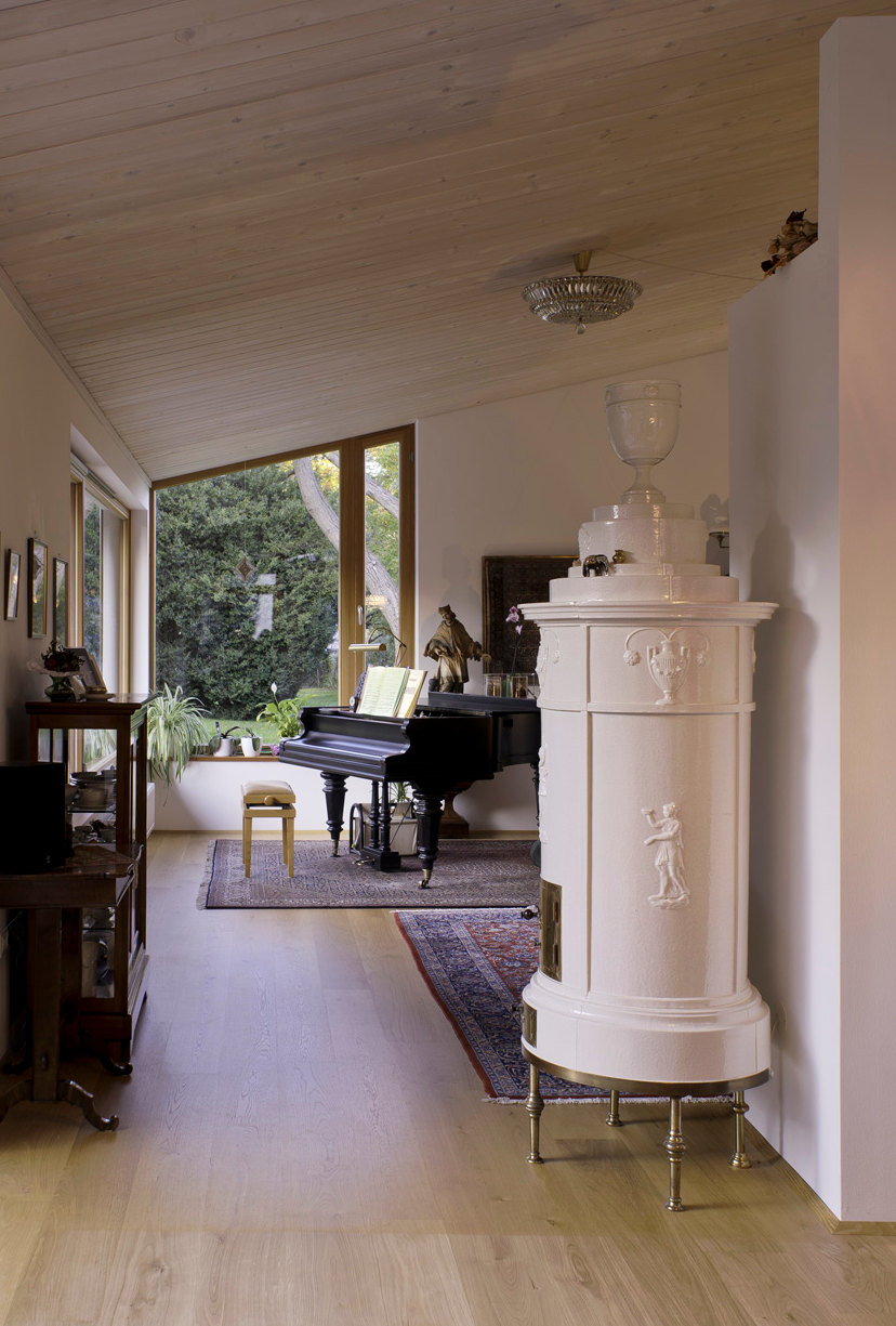 Living room with a large window, a grand piano, a small stool, a white ceramic stove with decorative reliefs, and various potted plants.