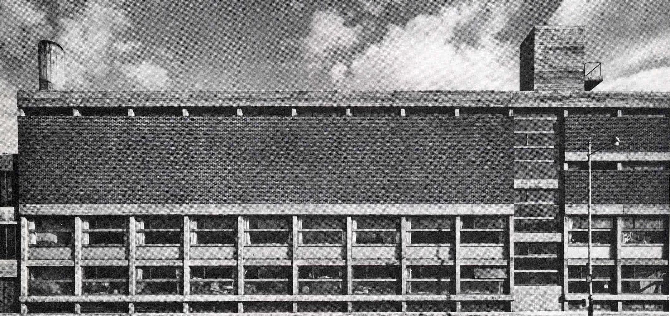 Black and white photo of a modern multi-story building with exposed concrete and large windows, set against a cloudy sky.