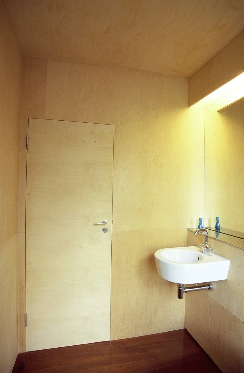 A minimalist bathroom with light wooden walls and ceiling, a small white wall-mounted sink, and a mirror above it. The door on the left matches the wood paneling, and there are two blue bottles on the shelf near the sink.