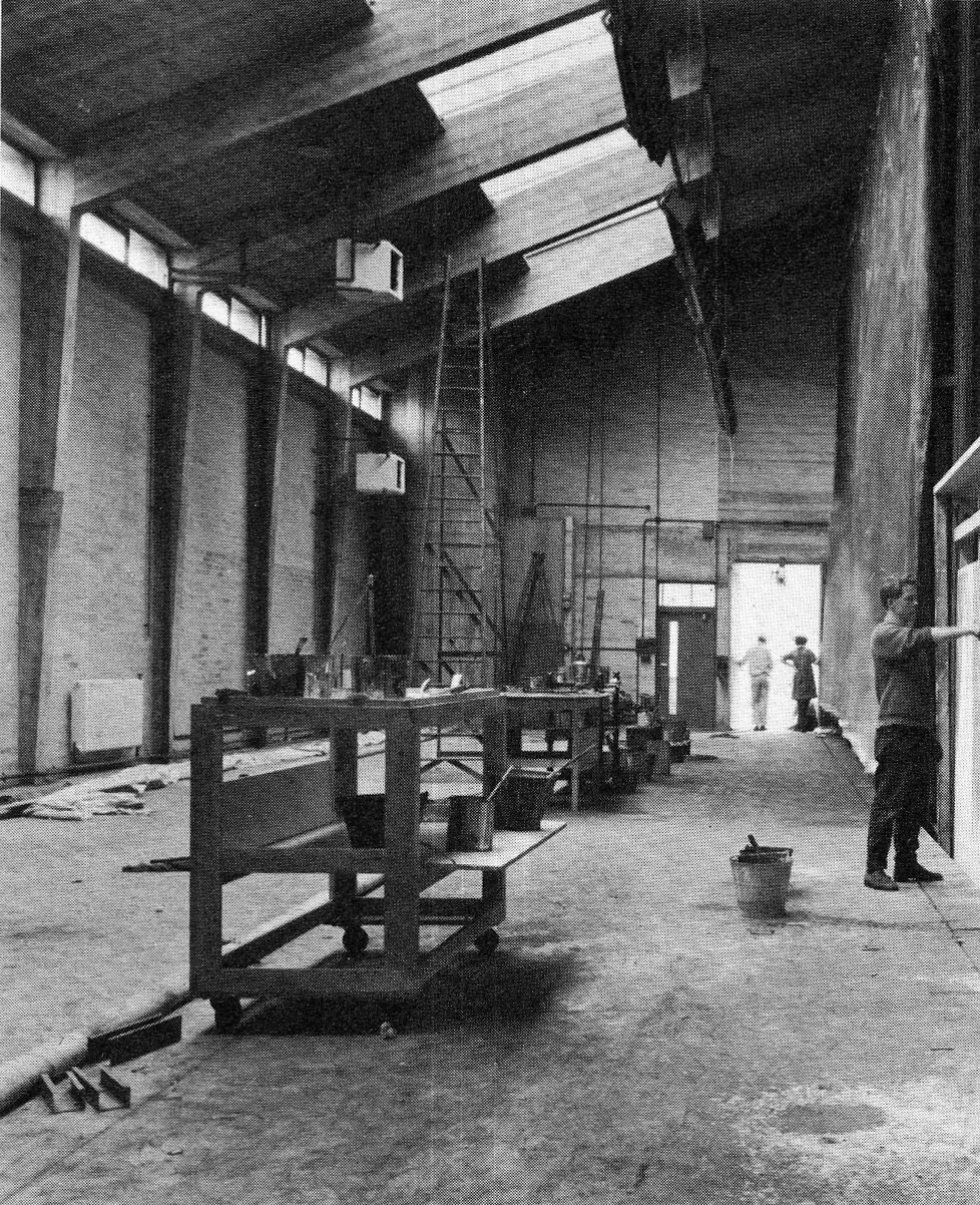 An indoor construction or renovation site with scaffolding, work tables, and workers. The space has high ceilings, exposed beams, and large windows near the ceiling, with some sunlight coming through. There are ladders, tools, and construction materials scattered around.