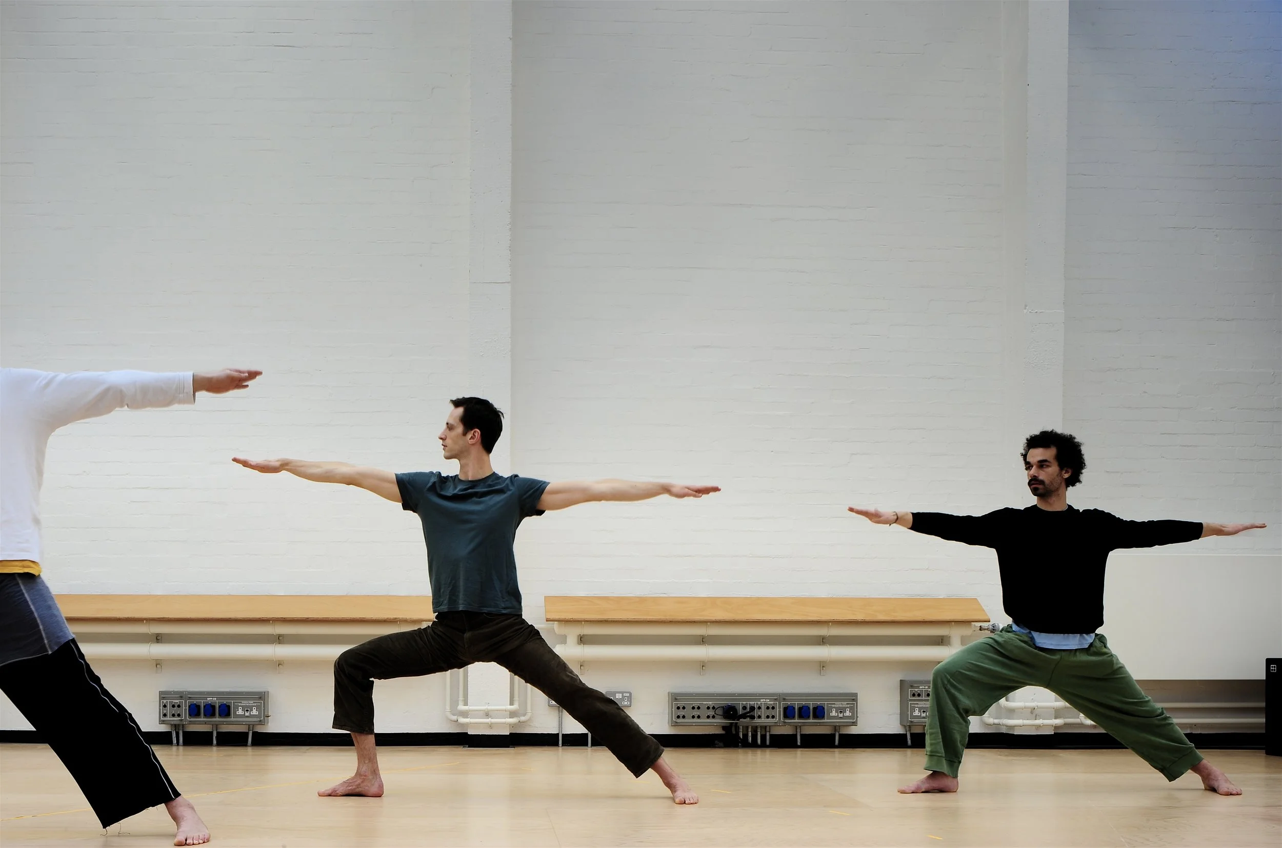 Three men practicing yoga in a studio, standing in warrior pose with arms extended and knees bent, wearing casual workout clothing.