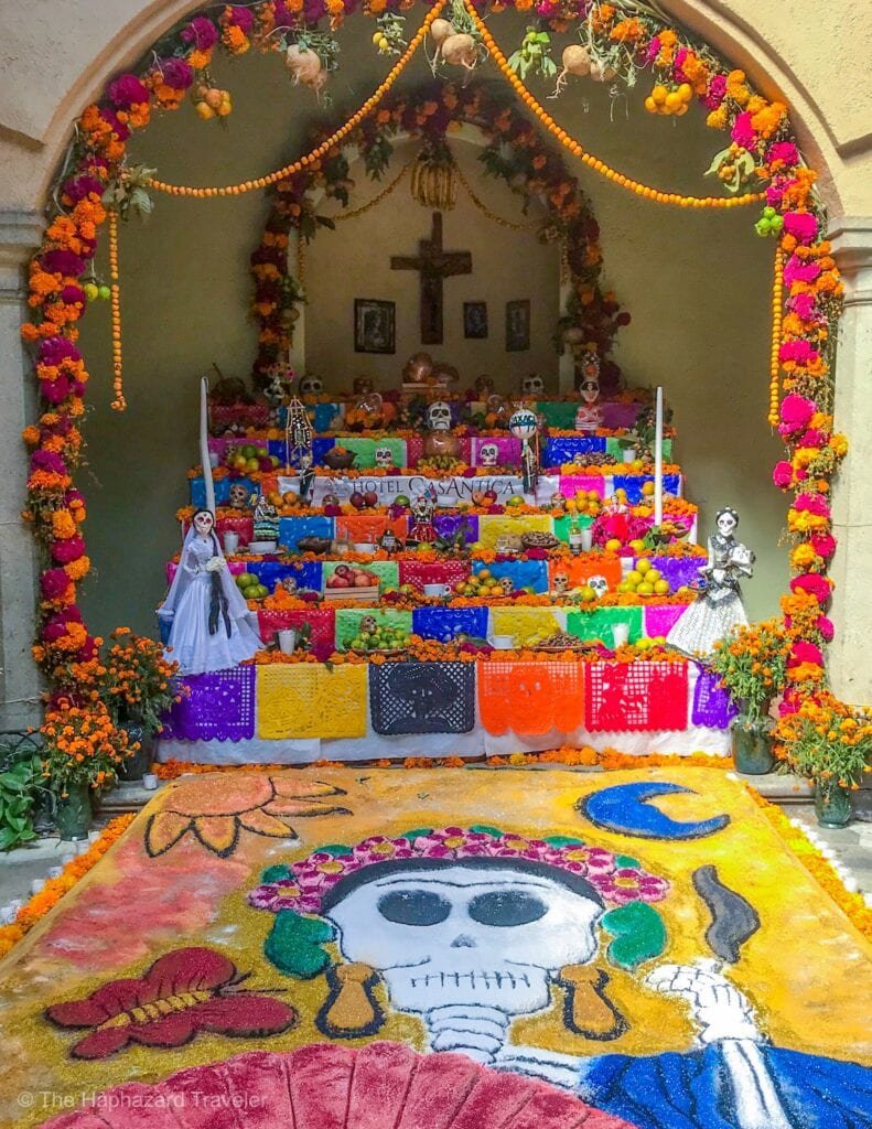 A colorful Frebana altar decorated with marigolds, papel picado, and various traditional items, commemorating Día de los Muertos. The altar features skull figures, candles, flowers, and photographs, with a prominent painted skull on the floor, a cross and framed pictures on the wall behind.