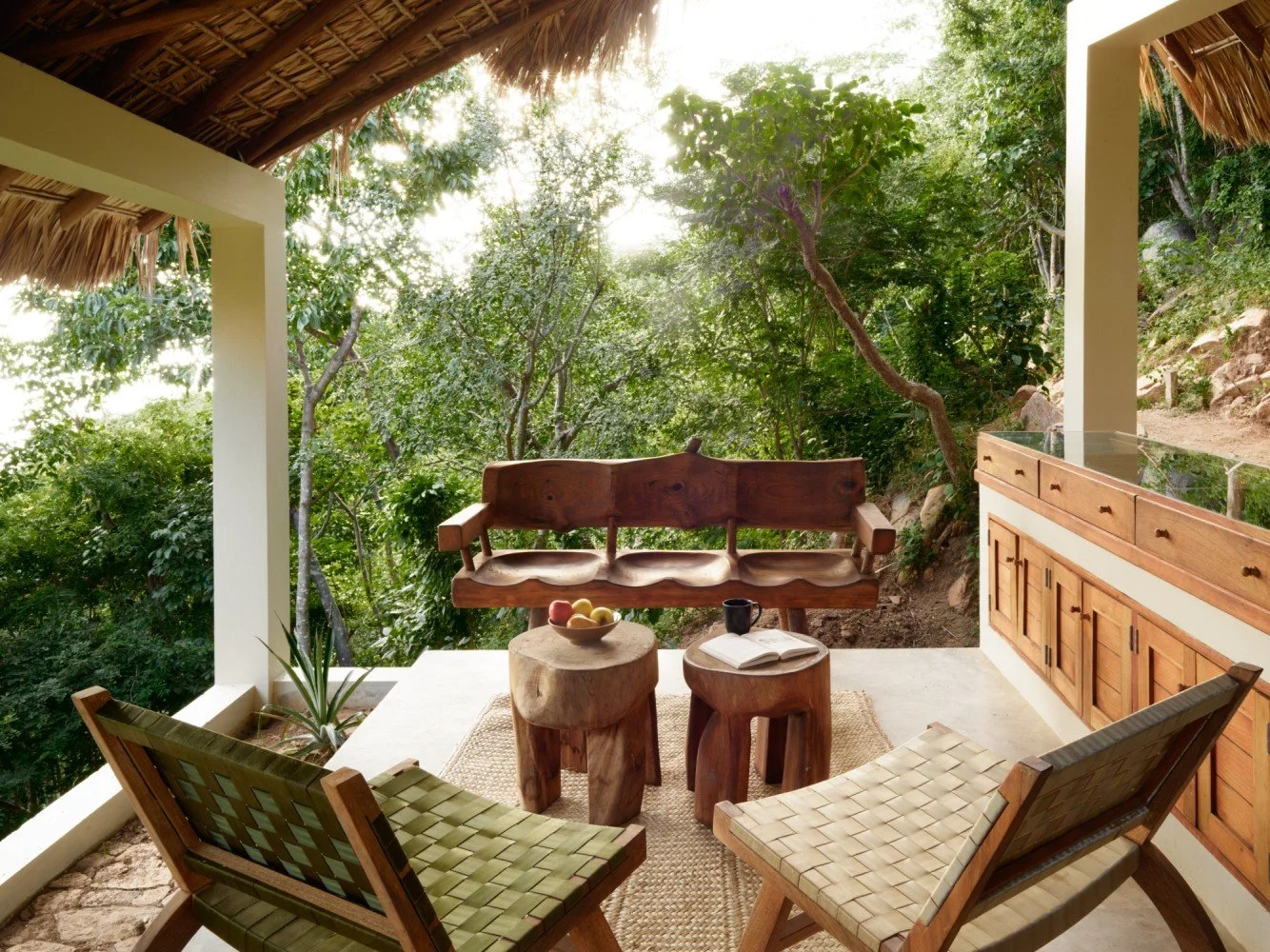 Outdoor patio area with wooden furniture and lush green trees in the background, sunlight shining through the trees.