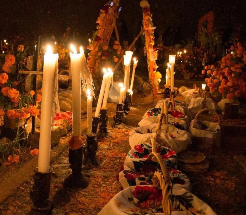 Nighttime altar with lit candles, flowers, and embroidered cloths on the ground, indicating a traditional celebration or memorial.