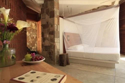 A bedroom with a bed covered by a mosquito net, a stone fireplace, a wooden table with flowers and fruit, and a window with blinds.