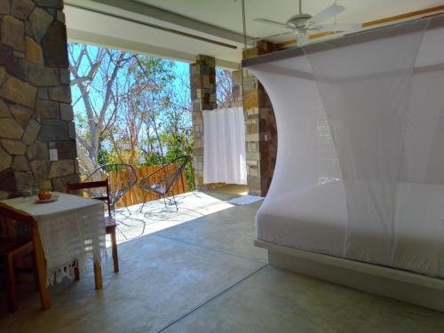 Sunlit indoor room with stone columns, a table with a tablecloth, chairs, a large white photo backdrop, and trees visible outside through large windows.