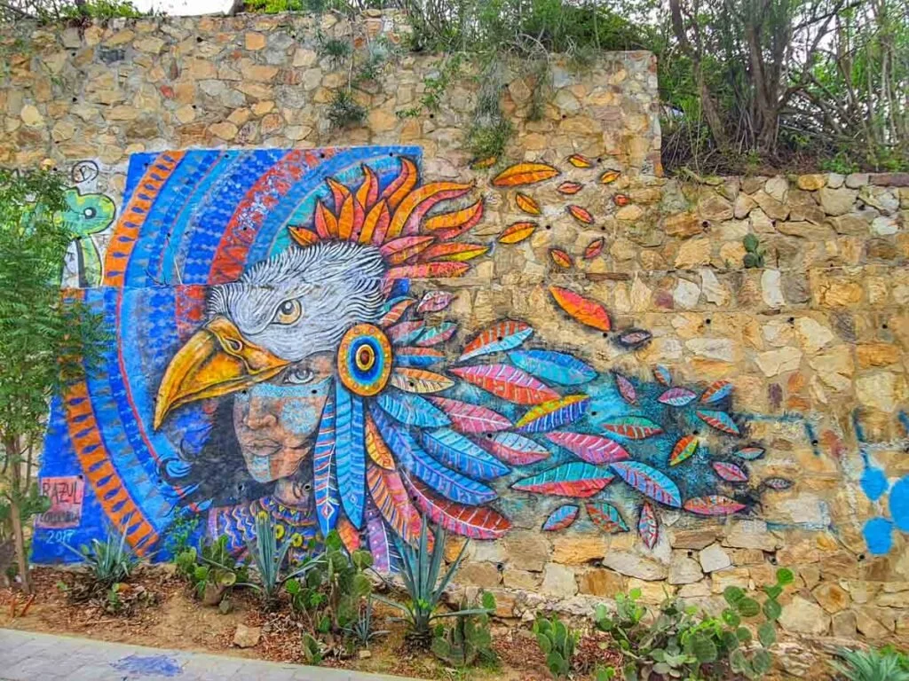 Colorful mural on a stone wall depicting a woman with feathers, a bird's head, and a child's face, with surrounding feathers and foliage.