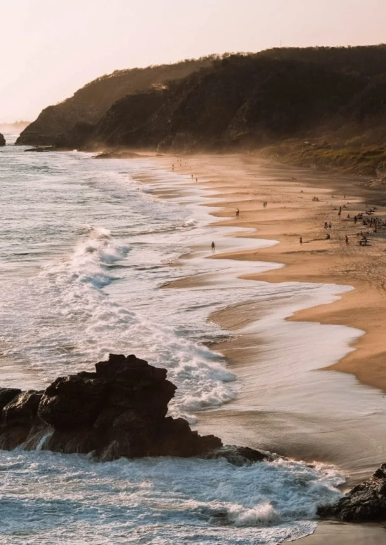 Aerial view of a sandy beach with waves crashing, rocky outcrop in foreground, and cliffs in the background during sunset.