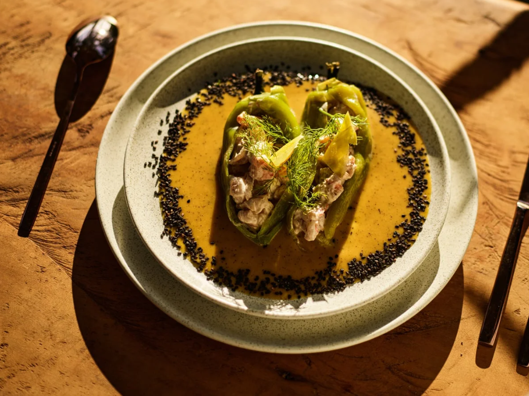 Green peppers stuffed with meat and vegetables, topped with herbs, in a yellow sauce, garnished with dill, served on a white plate with black sesame seeds around the edge, placed on a wooden table.