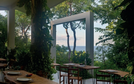 Indoor restaurant with large open window showing trees and ocean view, wooden tables and chairs, surrounded by lush greenery.