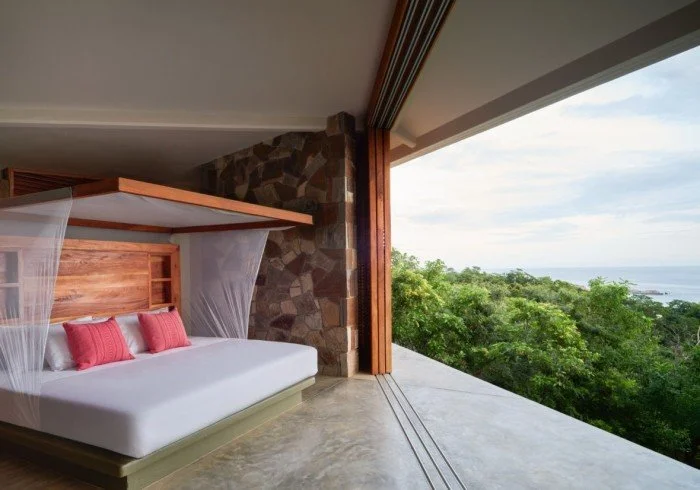 A bedroom with a large window overlooking greenery and the ocean, featuring a wooden bed with pink pillows and a canopy net.