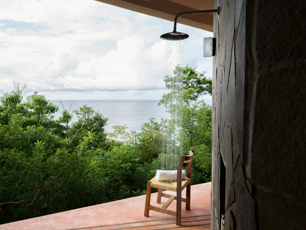 Outdoor shower with rain falling, overlooking lush green trees and a body of water in the distance, with a wooden chair and a rolled towel on it.