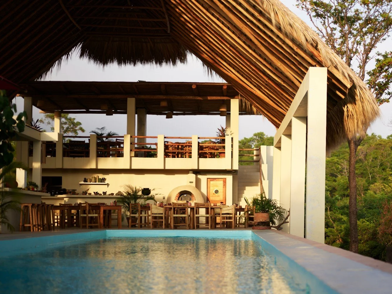 An outdoor pool with a thatched roof structure, tropical plants, and a seating area with wooden chairs and a table, overlooking a green landscape.