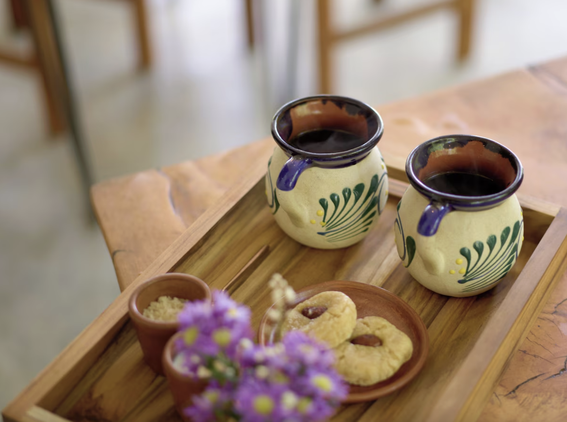 Two ceramic mugs of coffee on a wooden tray, with a small plate of cookies, a small bowl of sugar, and purple flowers in the foreground.