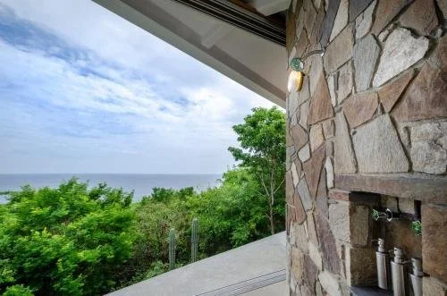 Outdoor space with a stone wall, string lights, and a view of green trees and the ocean.
