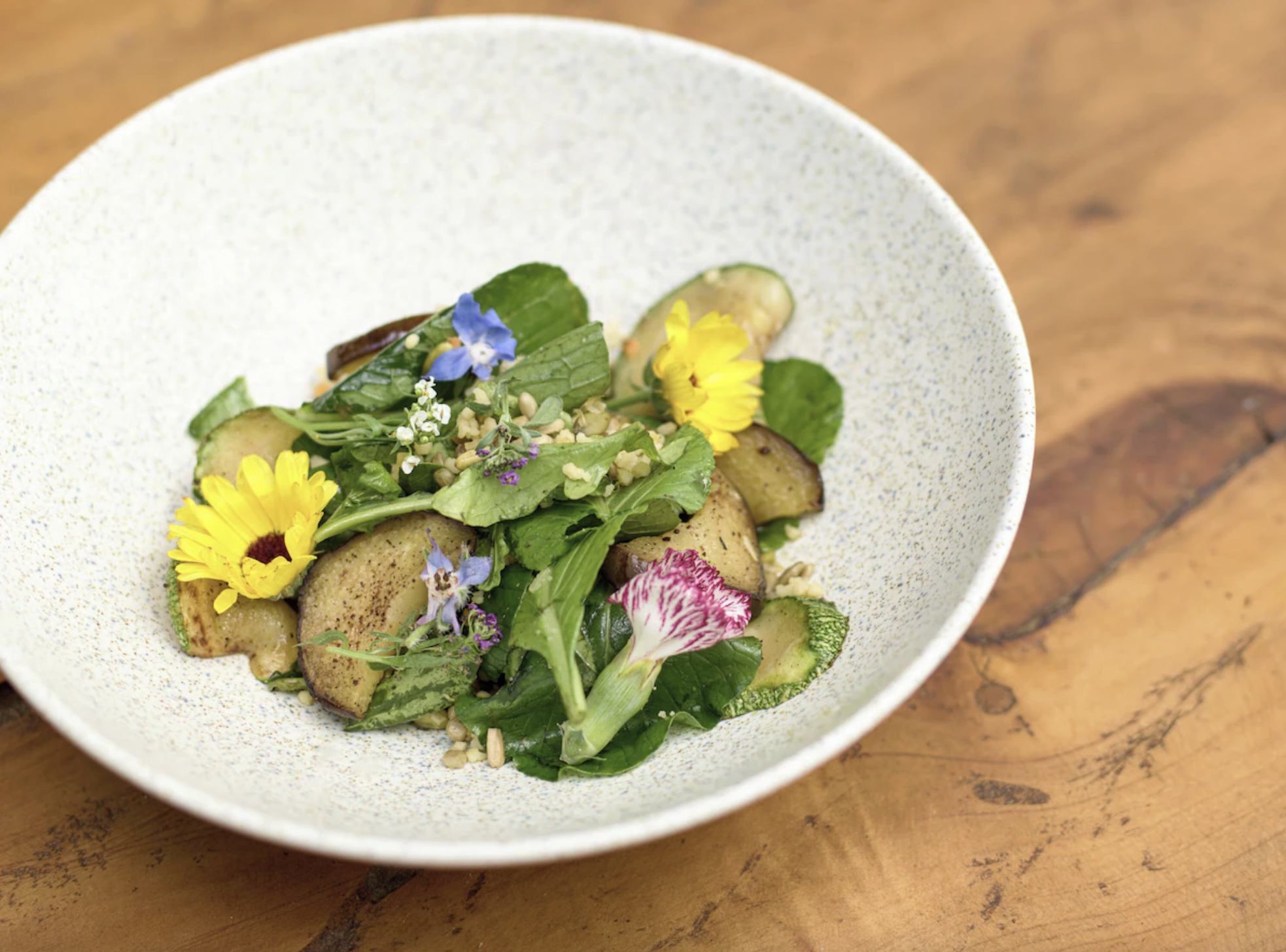 A salad with mixed greens, sliced eggplant, edible flowers, and seeds in a white speckled bowl on a wooden table.