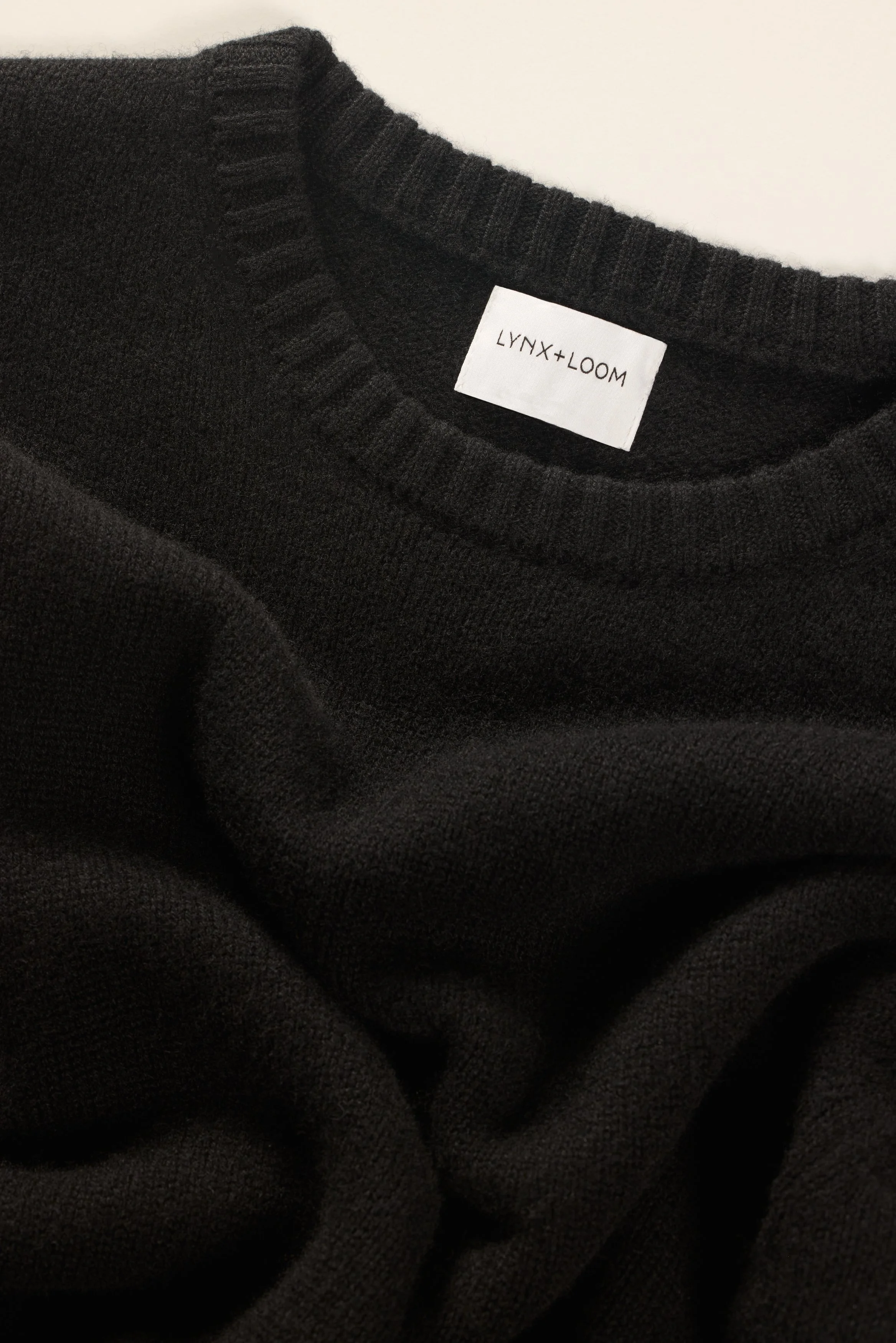 Close-up of a black Lynx + Loom sweater with a visible white tag, showing the brand name, laid on a white surface.