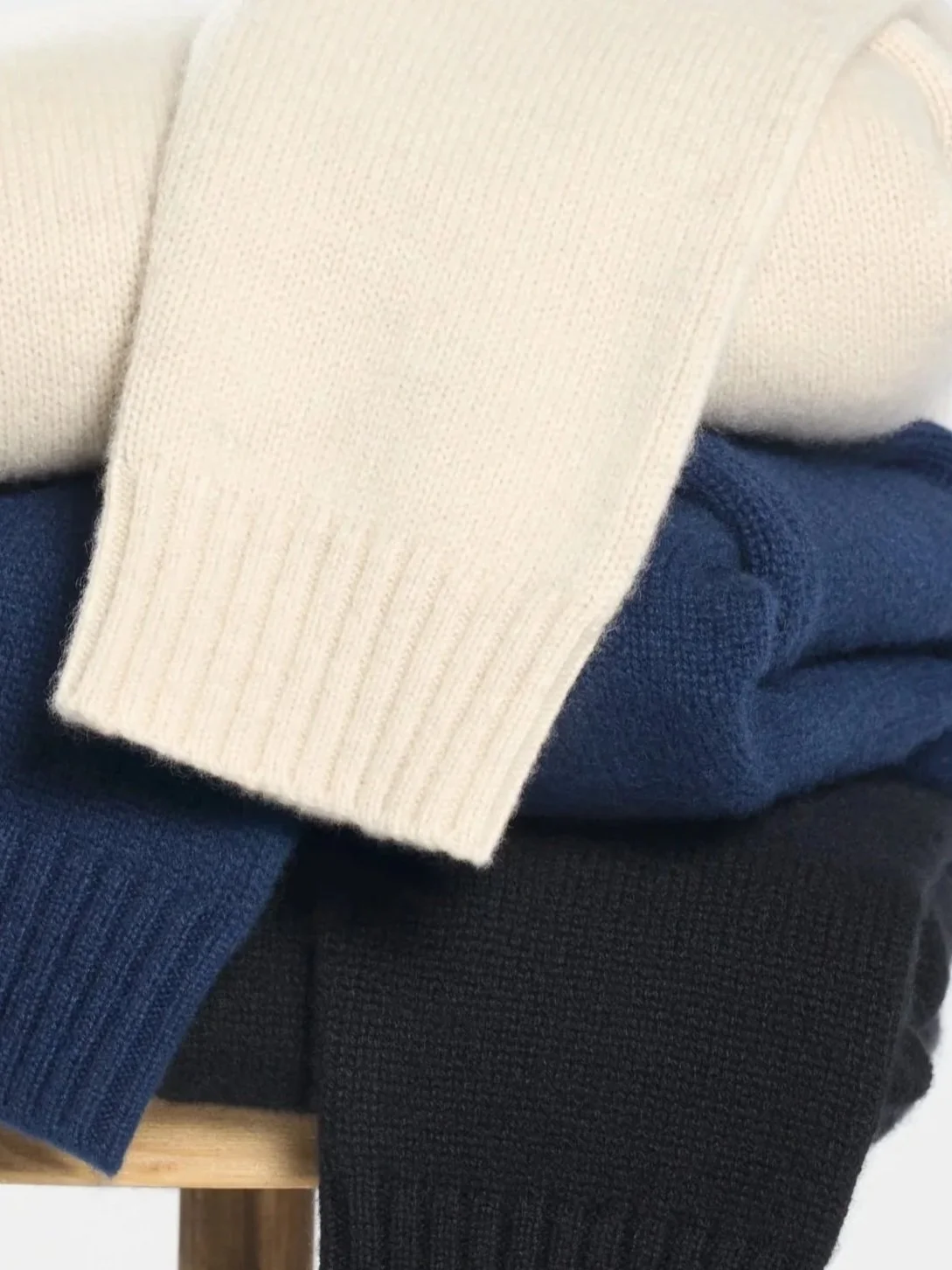Close-up of folded sweaters in beige, navy blue, and black, stacked on a wooden surface.