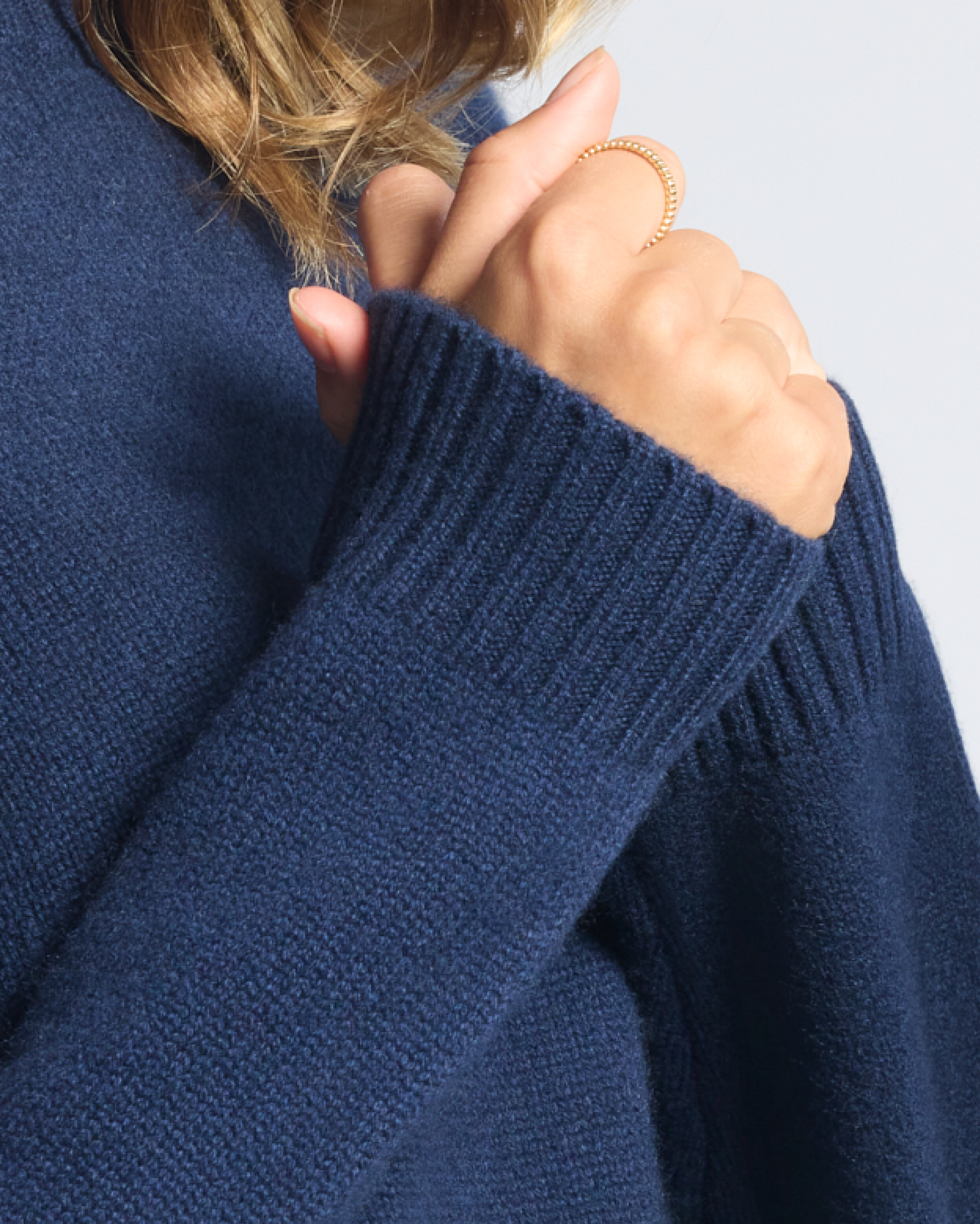 A person wearing a navy blue sweater with their hand near their face, showing a gold ring with small beads.