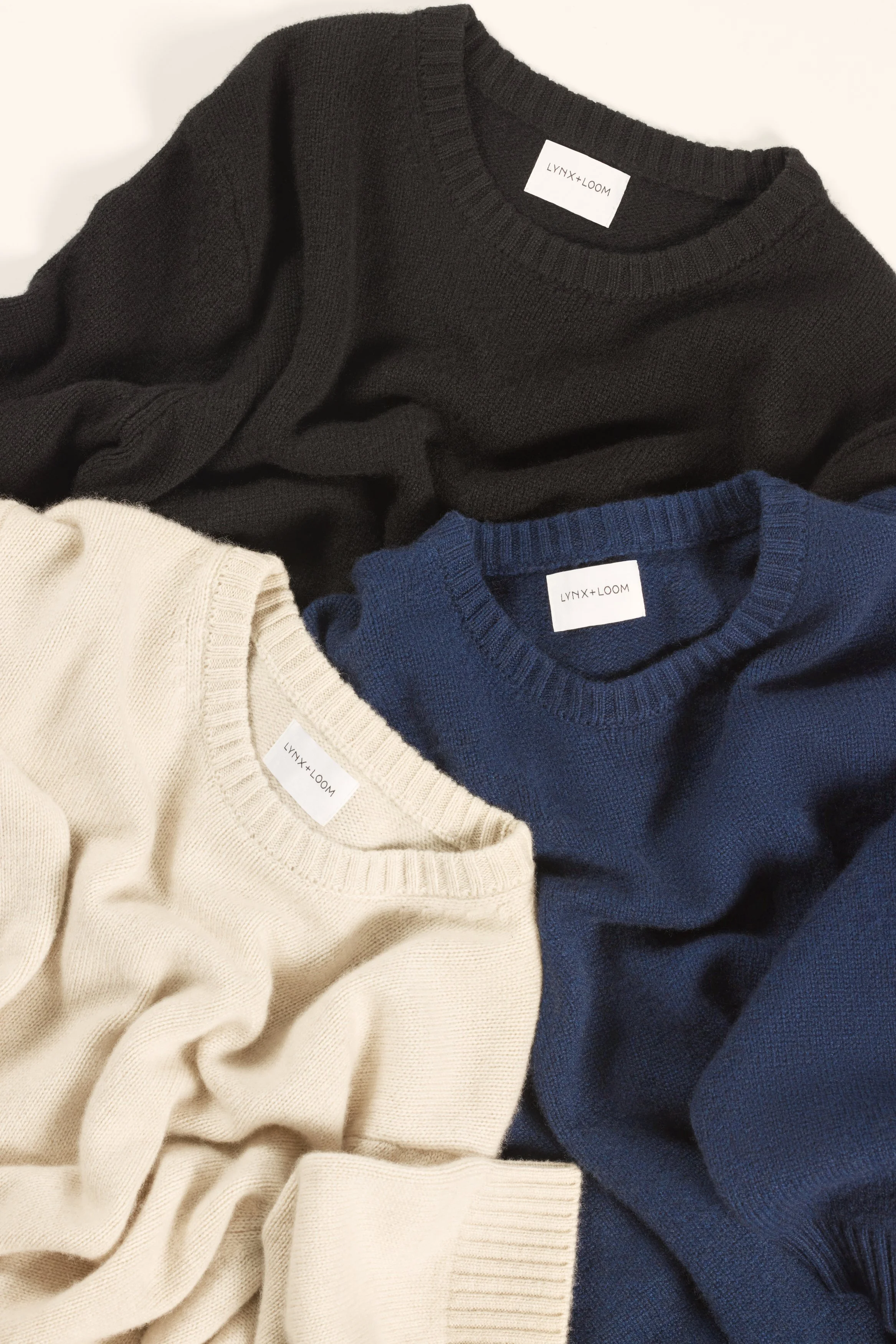 Three folded sweaters in black, blue, and cream, each with a white label from LYNN+LOOM, arranged on a white surface.