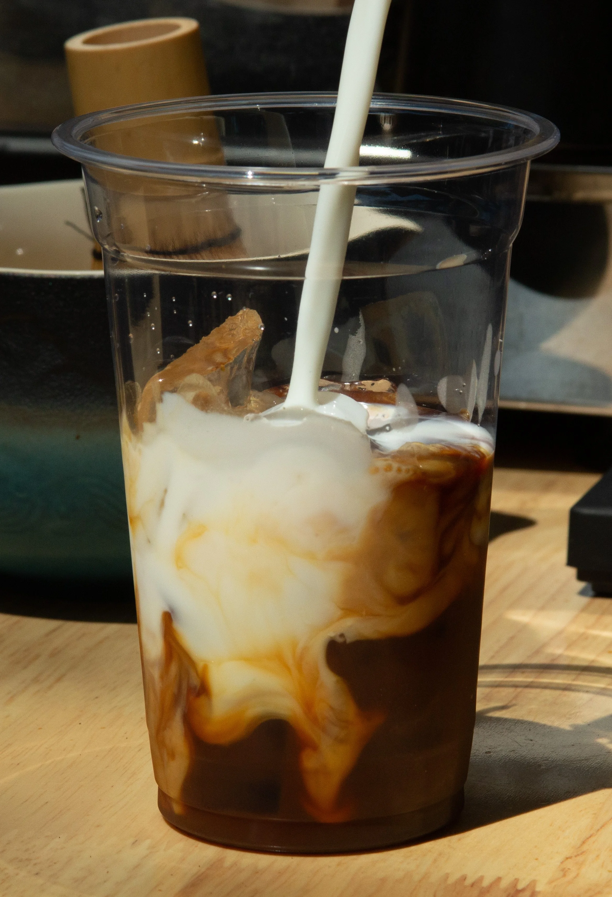 A tall clear plastic cup filled with iced coffee and milk, with a white straw inserted, placed on a wooden surface.