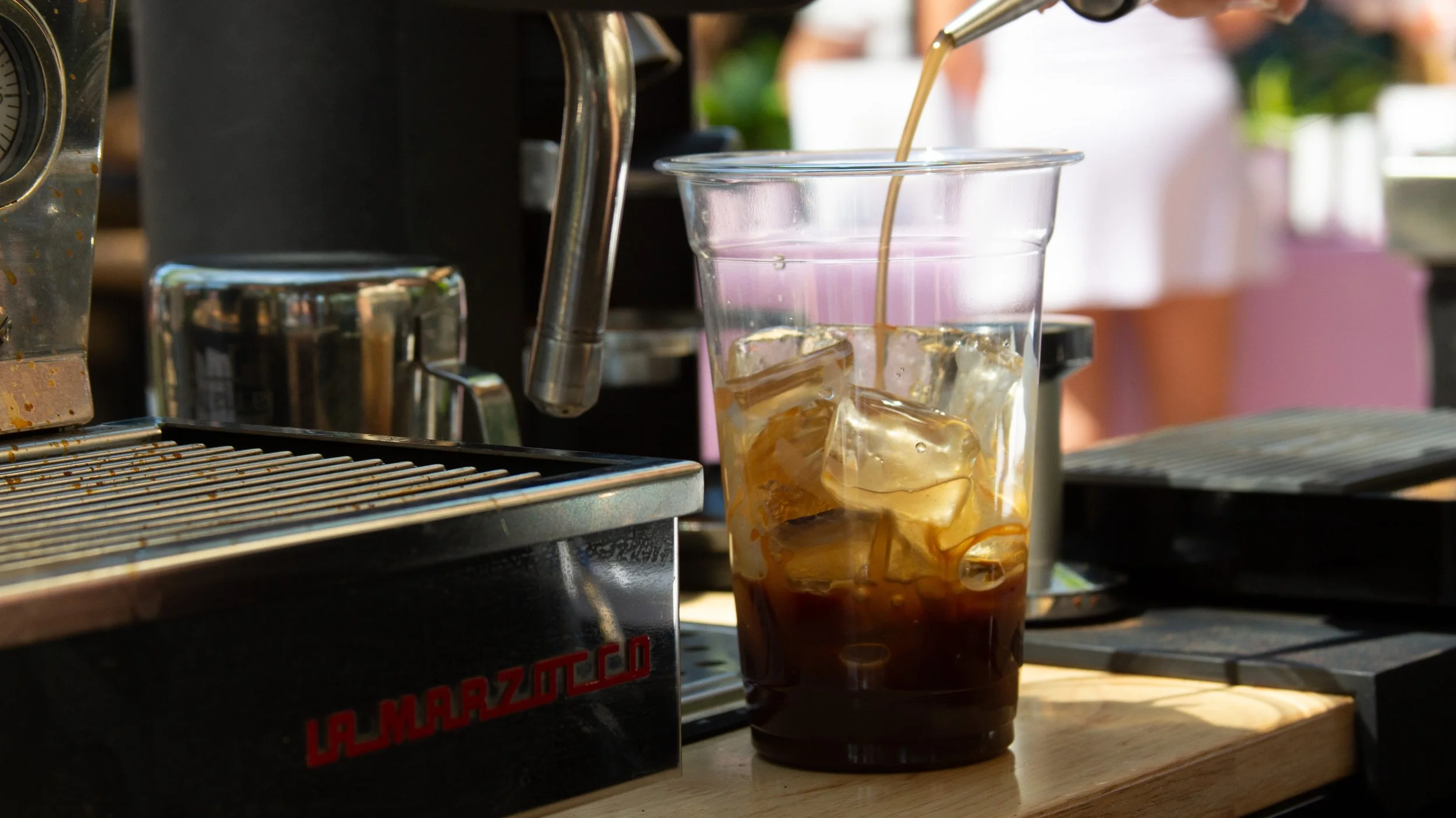 A tall glass of iced coffee with dark coffee at the bottom, ice cubes, and cream being poured in from a metal pitcher, set on a wooden surface with coffee equipment in the background.
