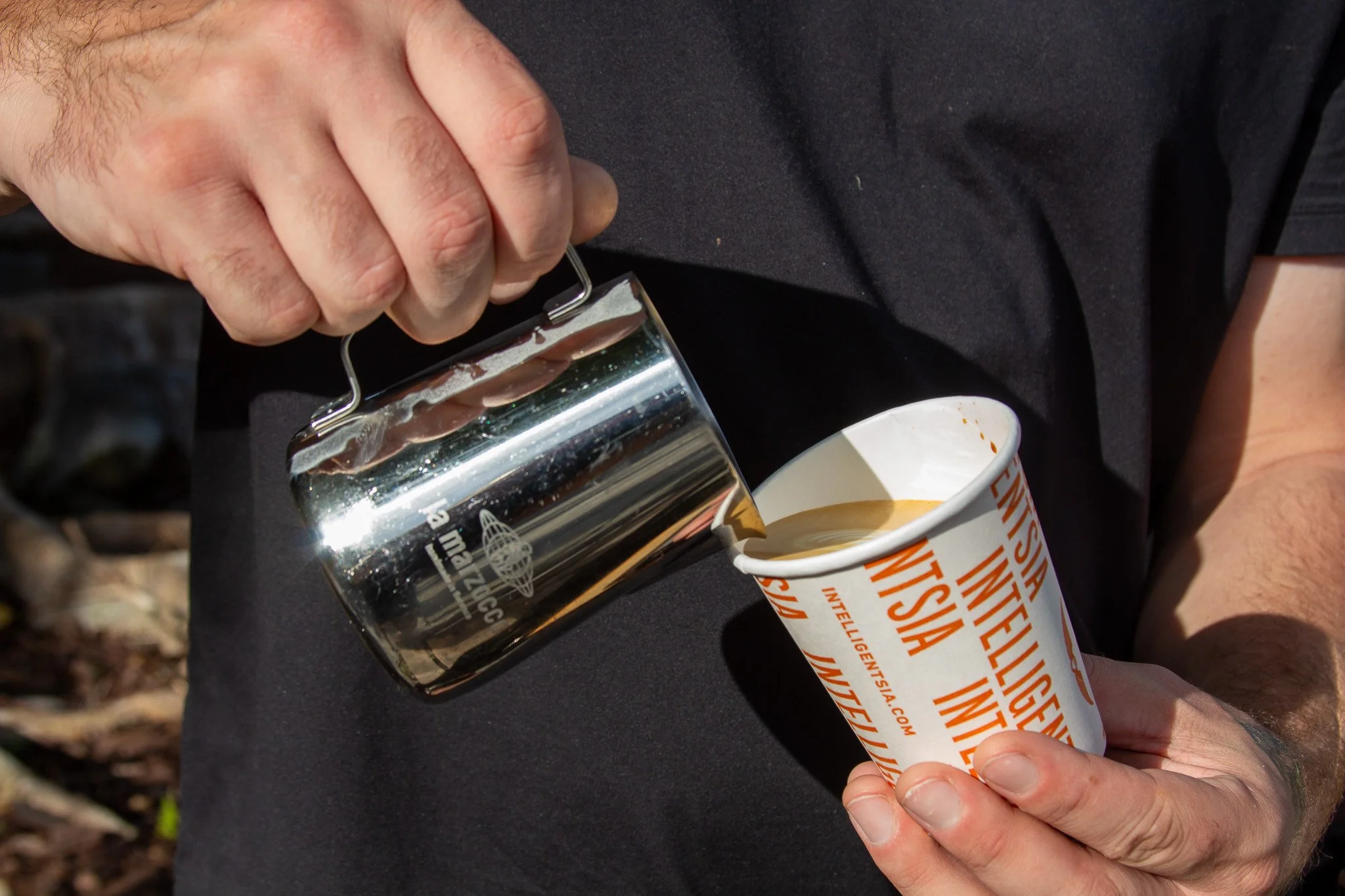 A person's hand opening a small glass jar containing a dark substance, with a disposable coffee cup nearby that has orange text and design on a white background.