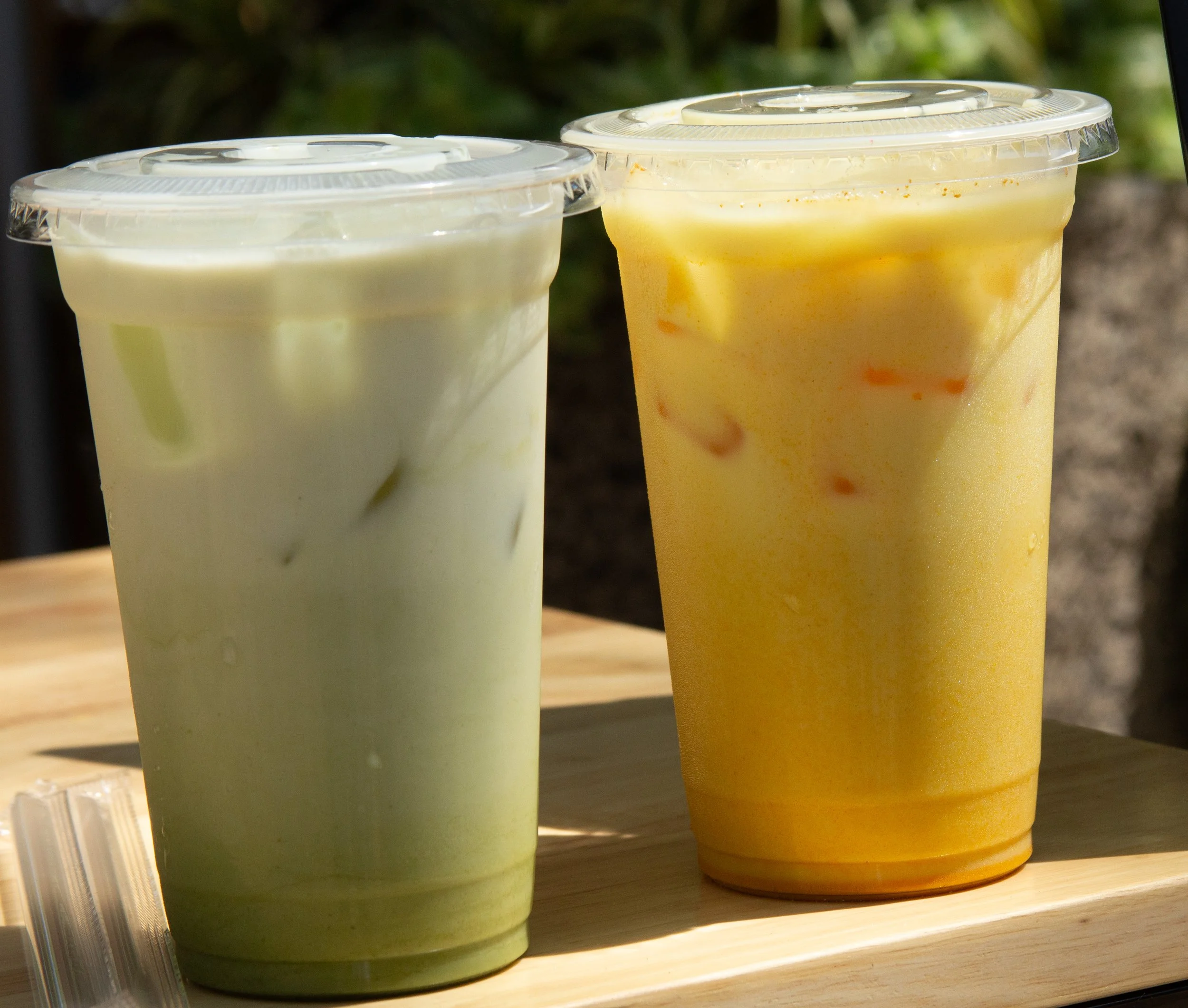 Two cups of creamy, colorful beverages on a wooden surface outdoors, one with a pale green color and the other with a yellow-orange hue with visible bits of fruit or garnish, covered with clear plastic lids.
