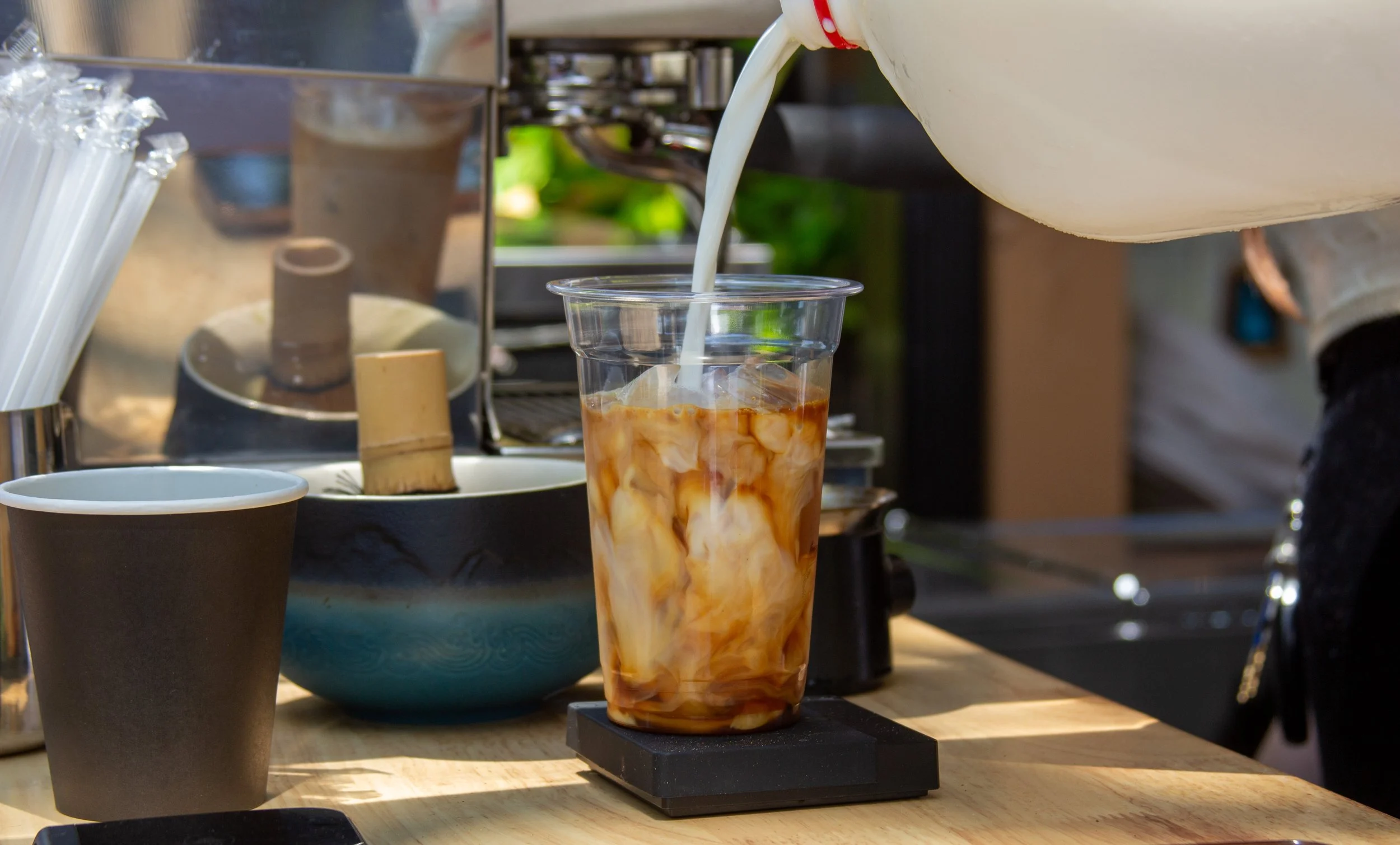 A coffee shop scene with a tall iced coffee beverage being poured with milk or cream, on a wooden counter. There are paper coffee cups, a bowl, and coffee-making equipment in the background.