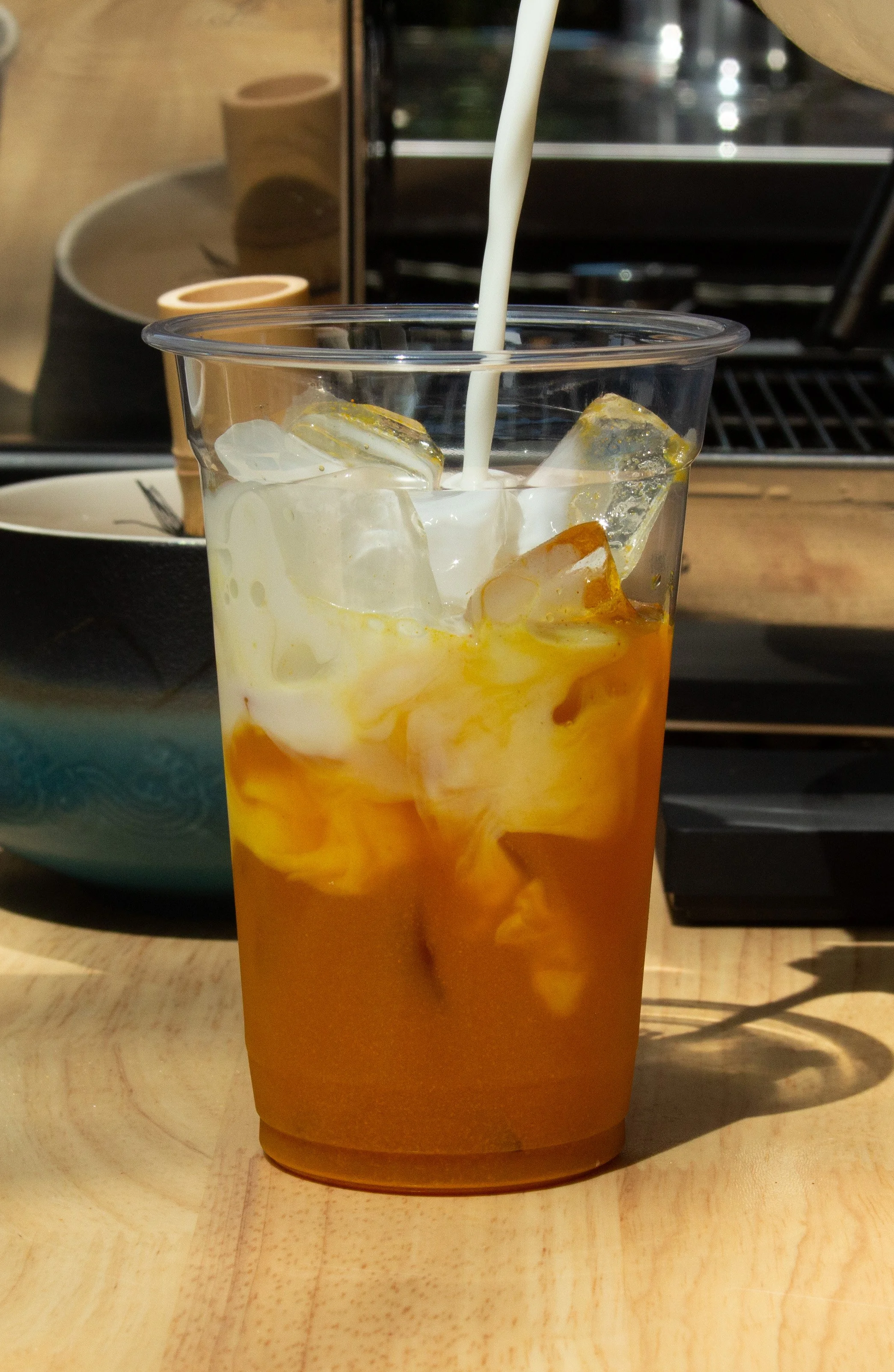 A tall clear plastic cup filled with ice, turmeric, and milk, with a stream of milk, placed on a light wooden table.