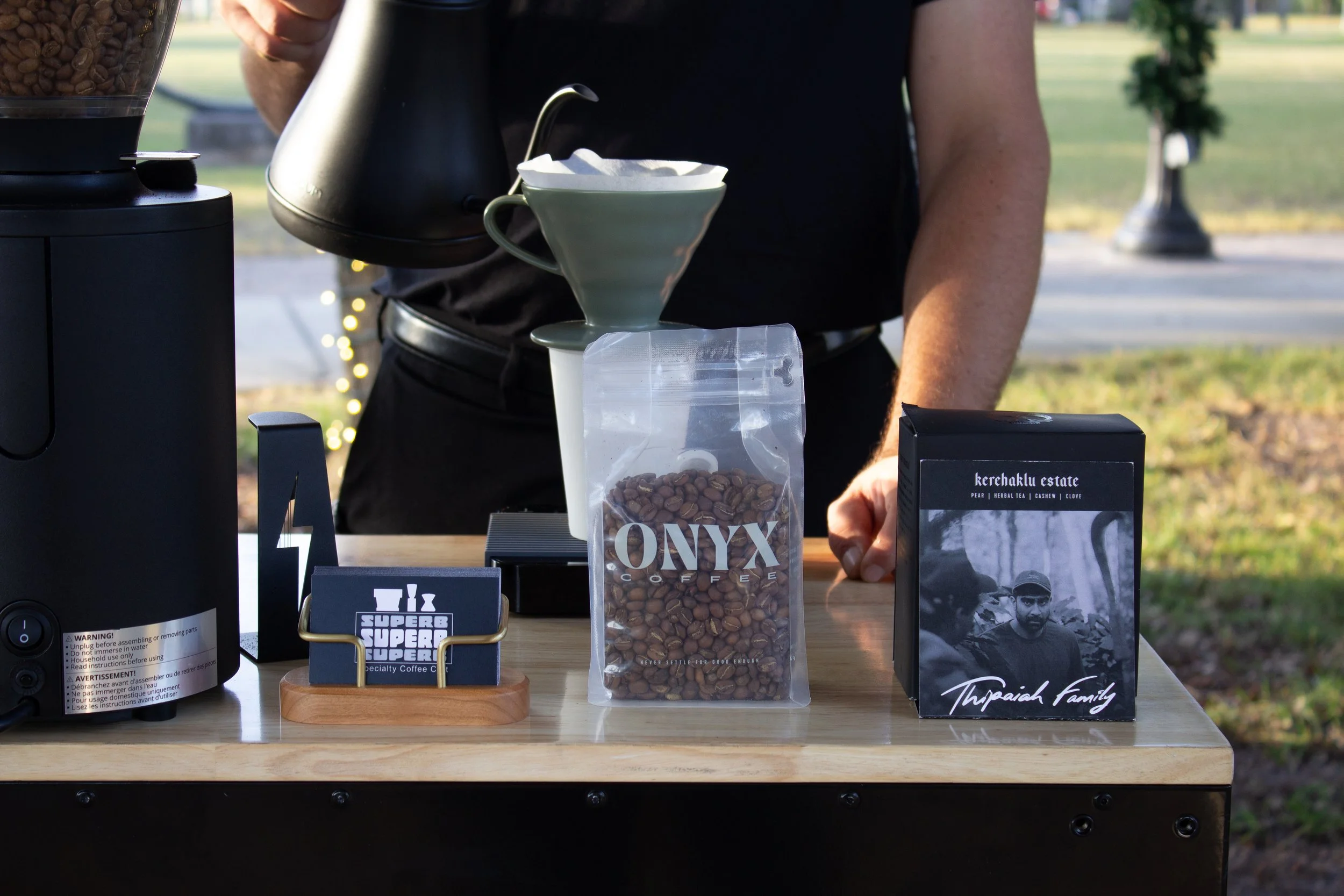 A person making pour-over coffee outdoors, with coffee beans, a coffee bag labeled 'ONYX Coffee,' and a black box on a wooden table.