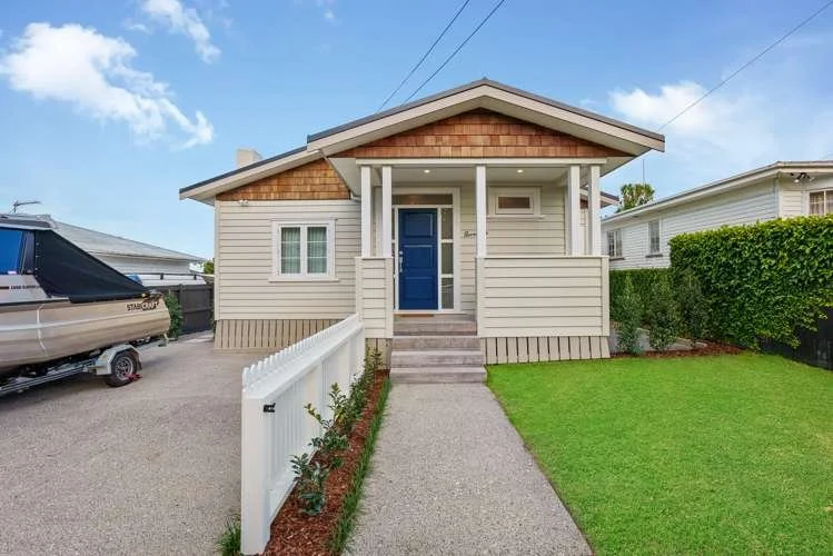 A small, single-story house with a blue front door, white siding, and a front porch with steps. There is a gravel driveway on the left with a boat on a trailer, and a green lawn with a white picket fence on the right. The sky is partly cloudy.