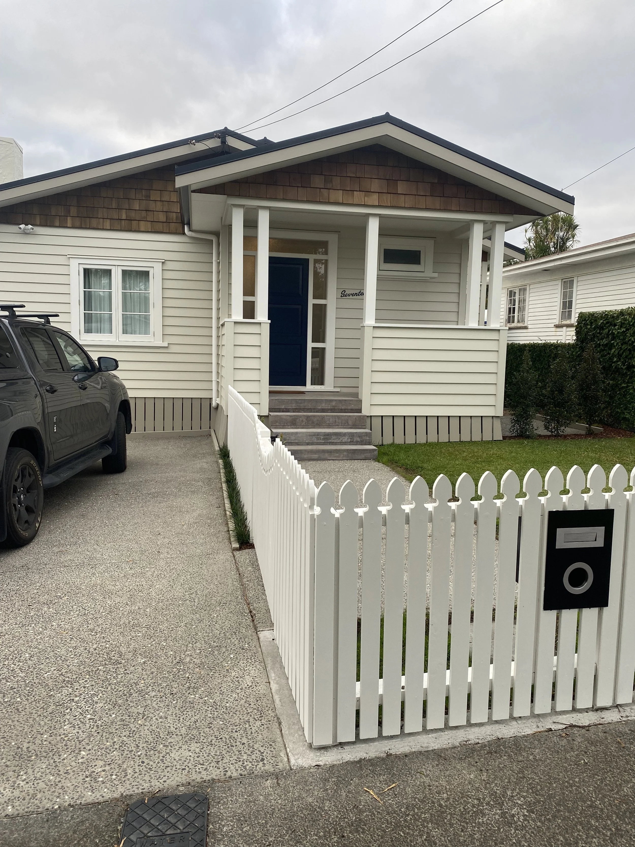 A white picket fence surrounds a small front yard with greenery. Behind it is a house with white siding, a blue front door, and a small porch with steps. There is a black car parked on the driveway to the left. The sky is cloudy.