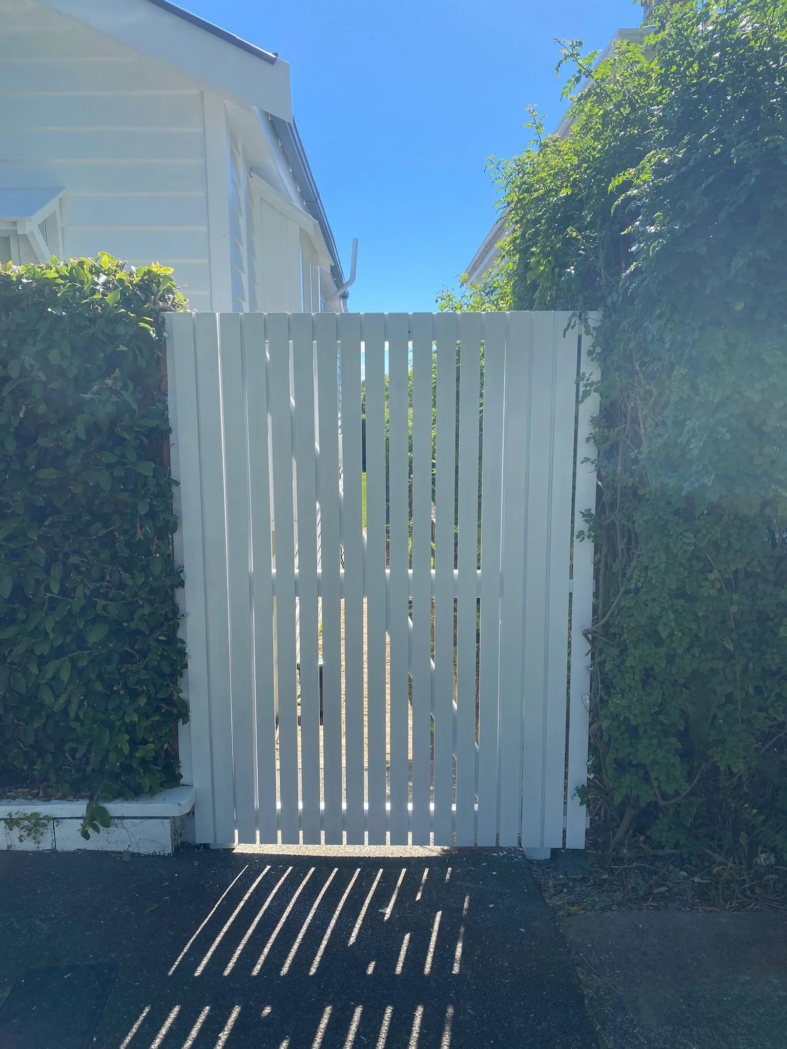White wooden gate with vertical slats between green bushes on both sides, sunlight casting shadow patterns on the ground, part of a white house visible on the left, and a clear blue sky.