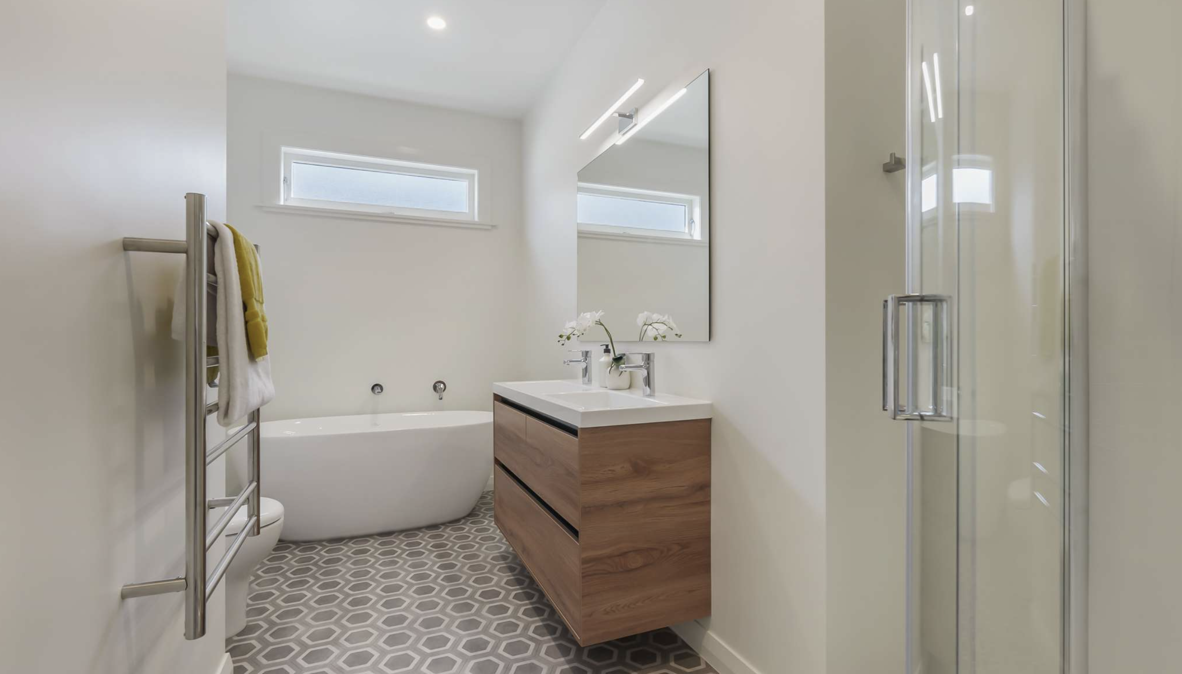 Modern bathroom with white walls, hexagon patterned gray floor, bathtub, wooden vanity with two sinks, large mirror, towel warmer, and glass shower enclosure.