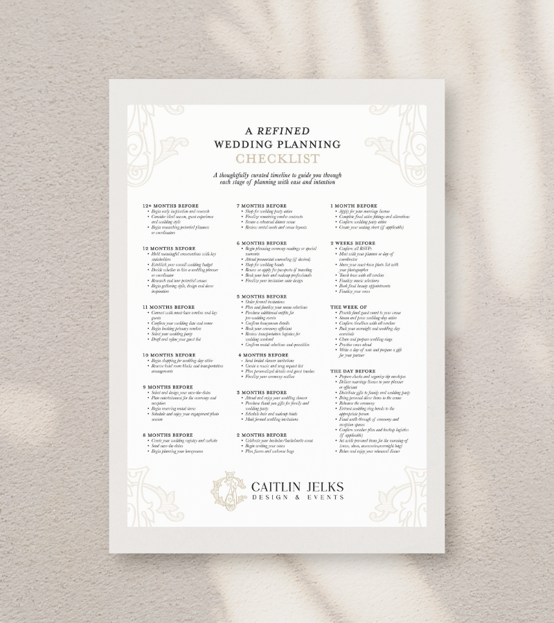 A wedding planning checklist on a white paper with decorative floral elements at the corners, placed on a beige textured surface. The checklist includes timelines and tasks for planning a wedding, from 12 months before to the week of the event, with Caitlin Jelks Design & Events branding at the bottom.