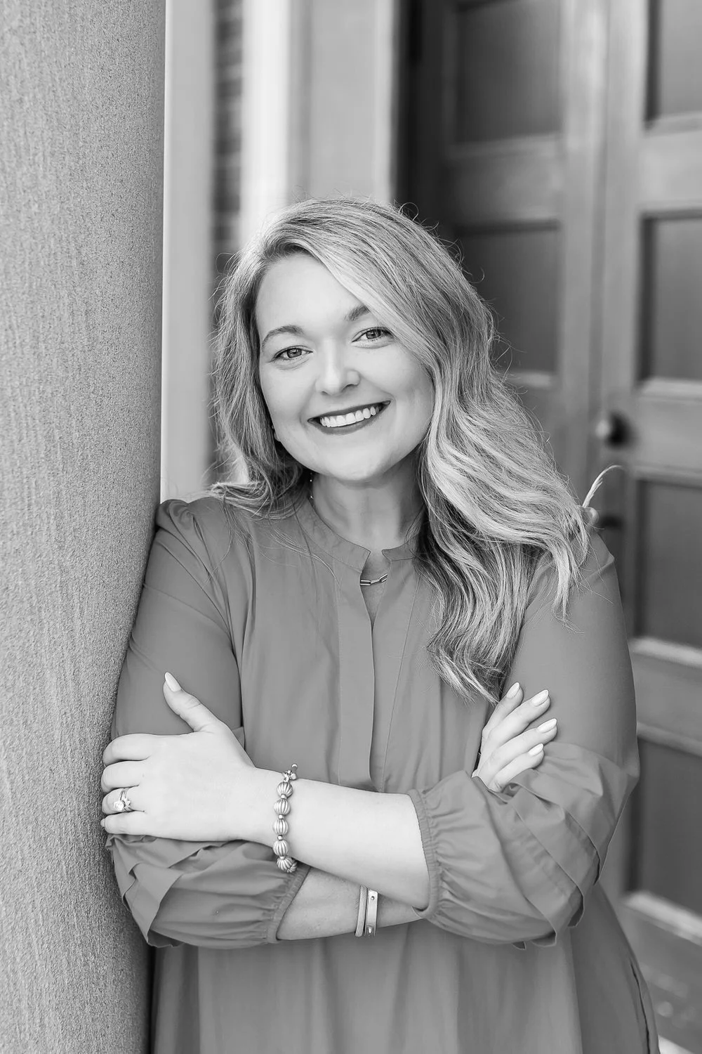 Black and white portrait of a smiling woman with wavy hair, crossed arms, wearing a long-sleeve blouse, standing outdoors against a wall with a wooden door in the background.