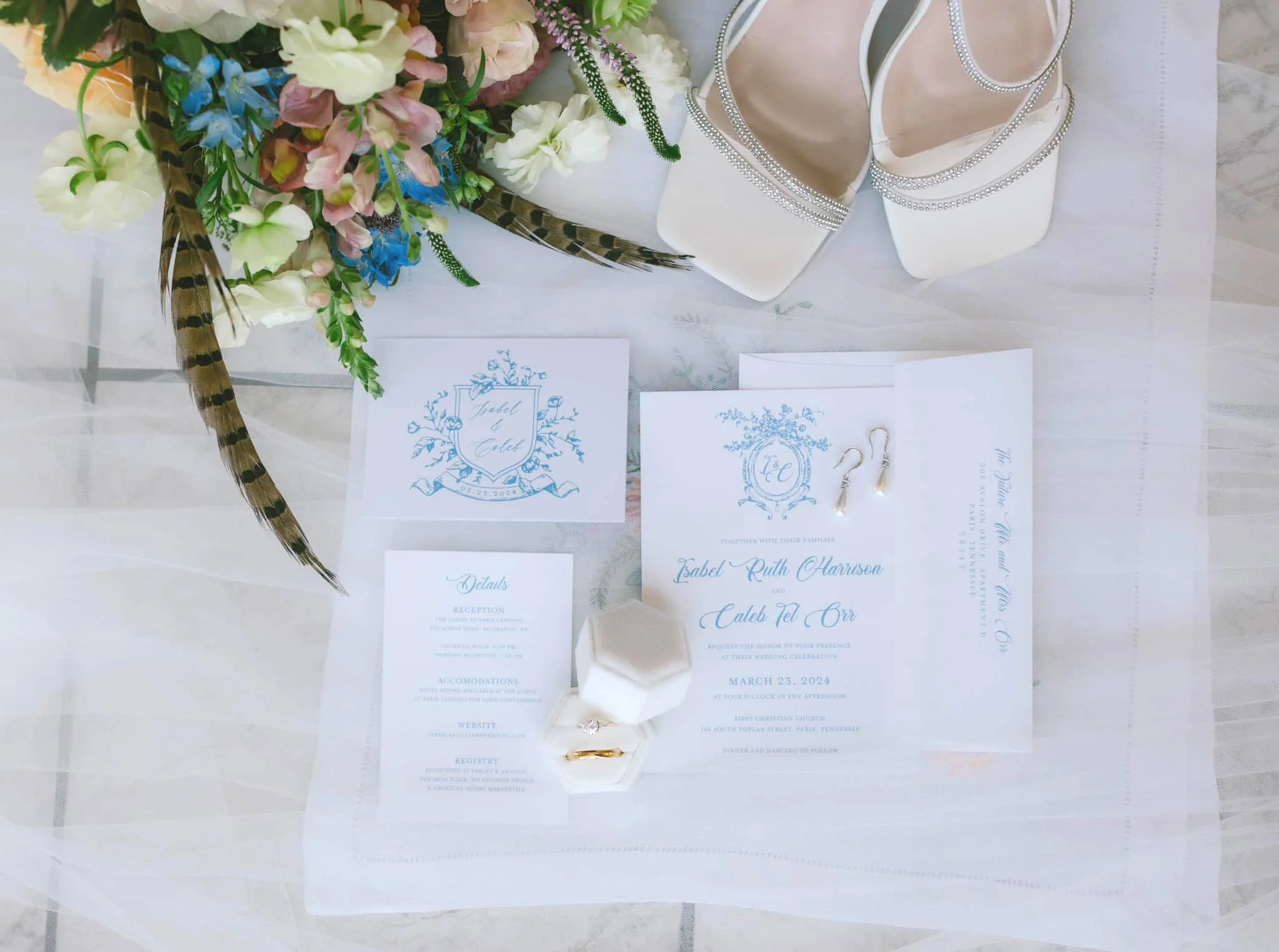 Wedding invitation suite with blue floral designs, a bouquet of mixed flowers, and a pair of white high-heeled shoes with rhinestone straps.