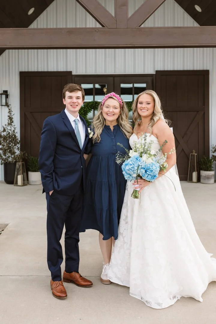 A wedding scene featuring a bride in a white wedding dress holding a bouquet of blue flowers, standing next to a groom in a navy blue suit, with a woman in a dark blue dress in the middle, all smiling outdoors in front of a barn-style building.