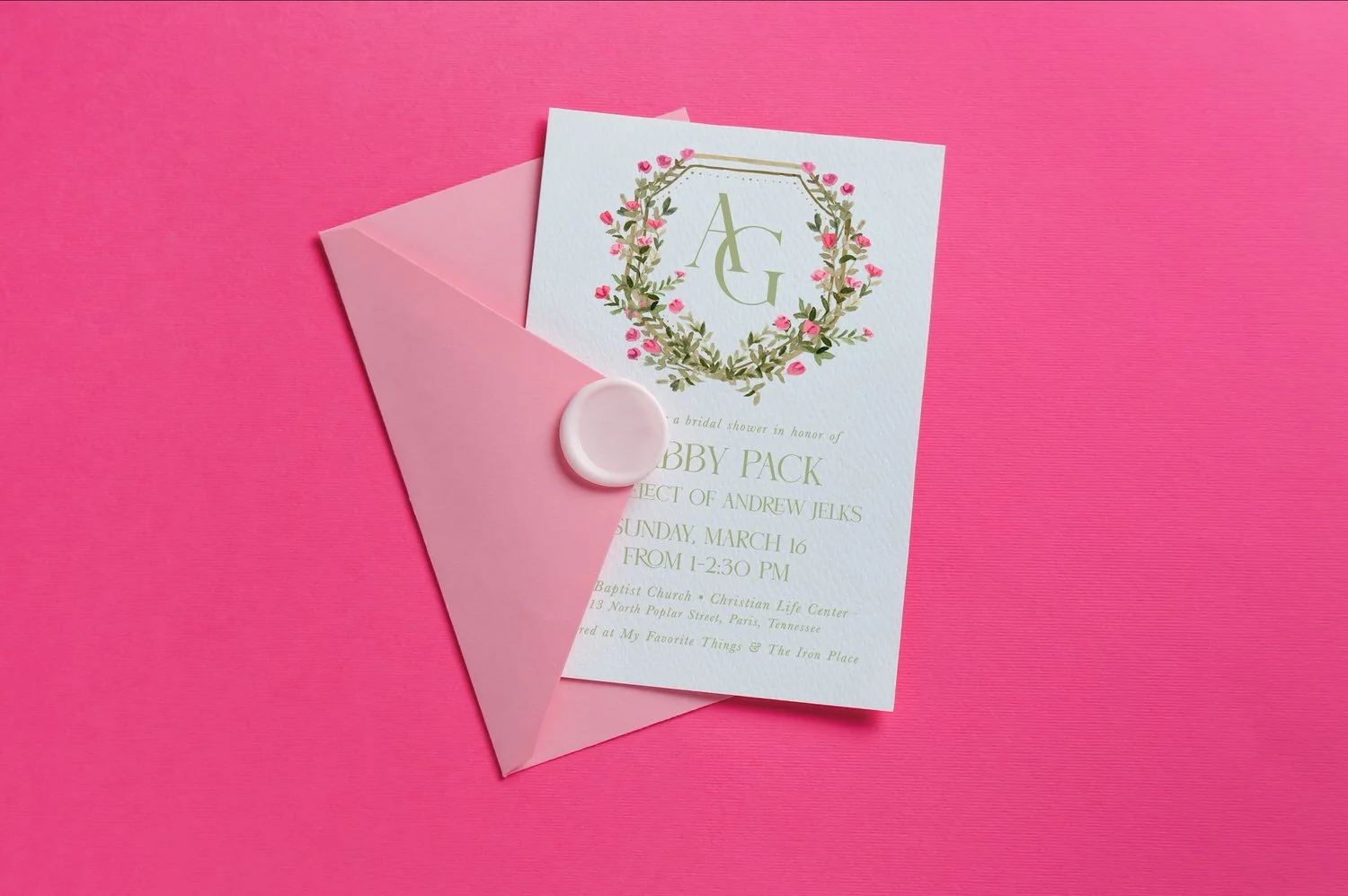 Invitation card for a bridal shower with a monogram 'A G' surrounded by a floral wreath, partially inside a pink envelope with a wax seal, on a pink background.