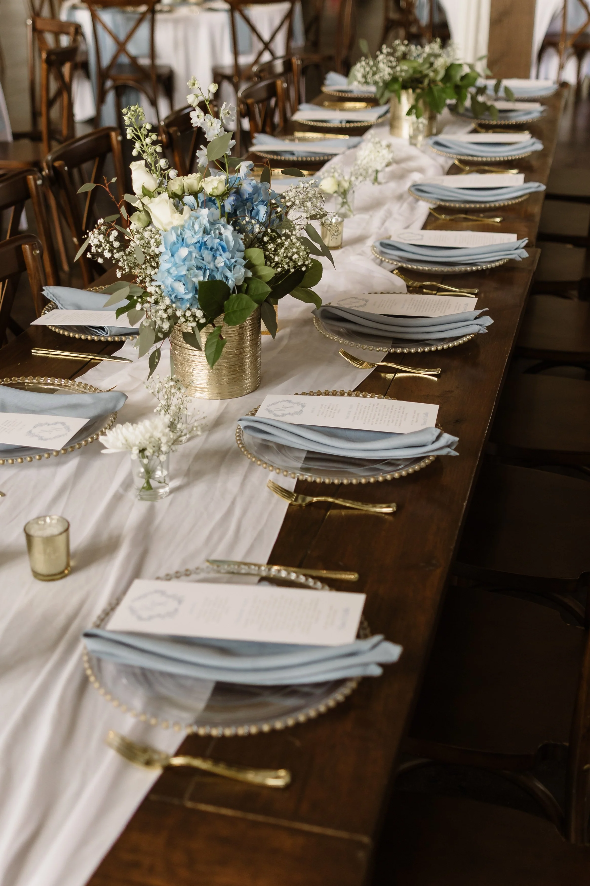 A long dining table decorated with white table runners, gold-rimmed plates, gold utensils, folded blue napkins, and floral centerpieces with white and blue flowers in gold vases, set for a formal event or wedding reception.
