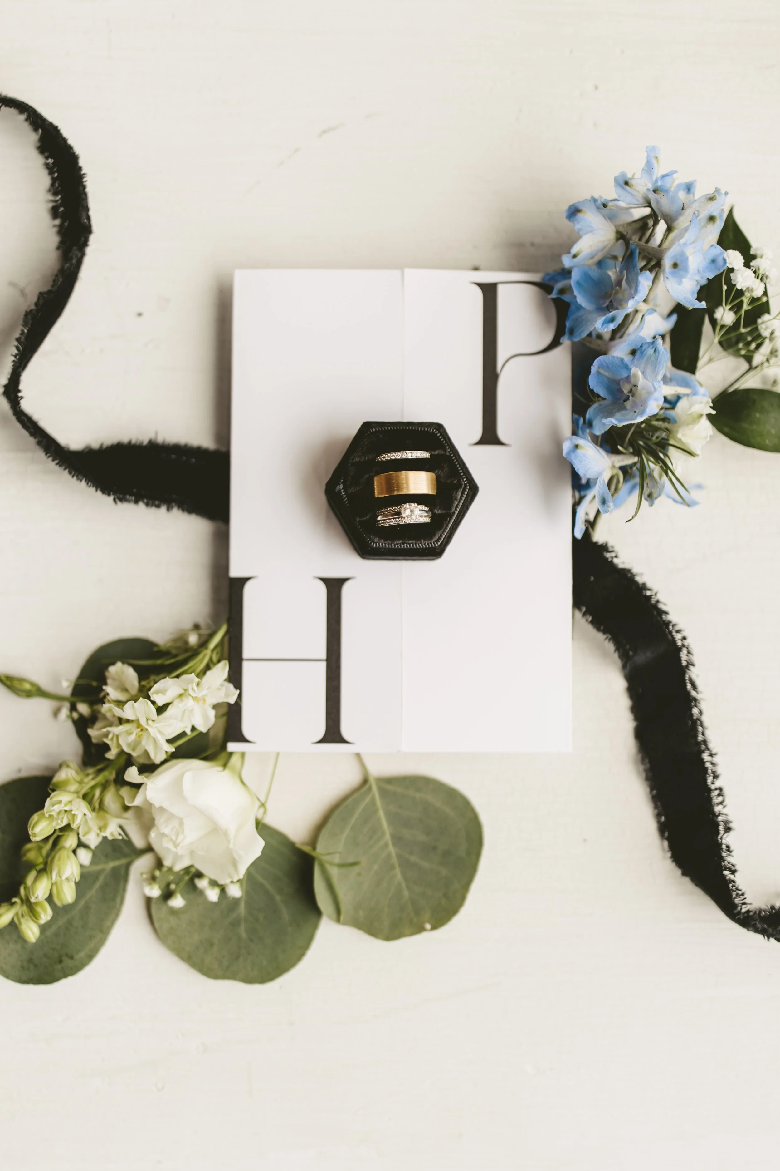 Wedding rings in a black ring box placed on a white photo book with black letters, surrounded by white and blue flowers and green leaves, on a white surface with black ribbon.