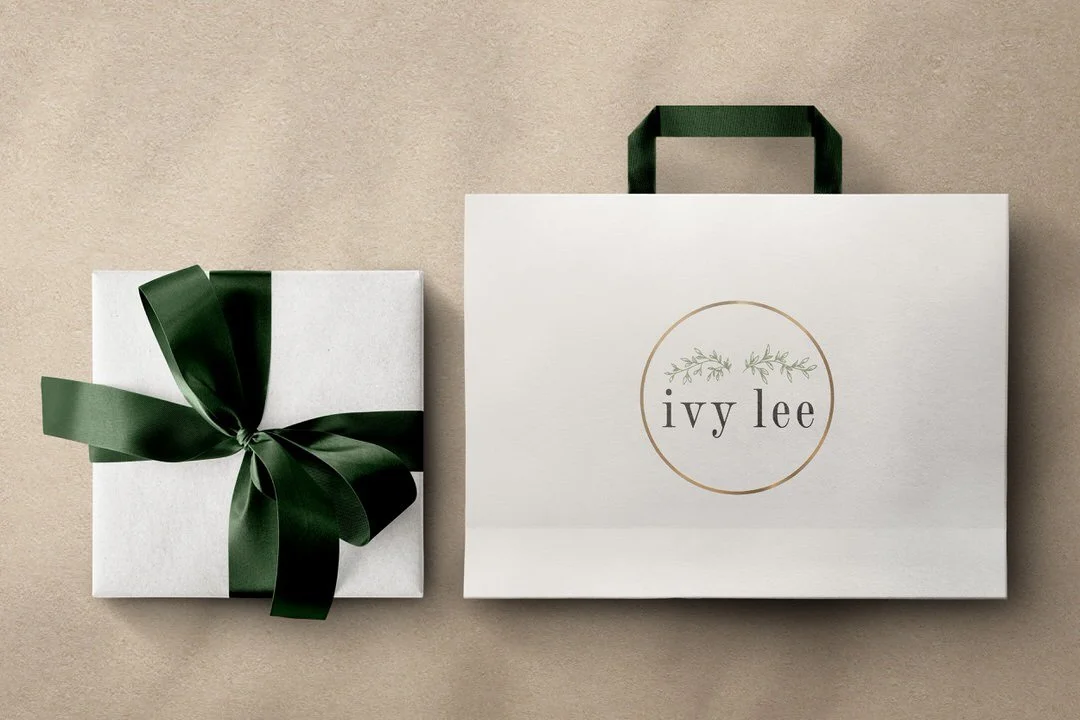 A wrapped gift box with a dark green ribbon and bow, and a white shopping bag with dark green handles and a circular logo reading 'ivy lee' in lowercase letters, on a beige background.