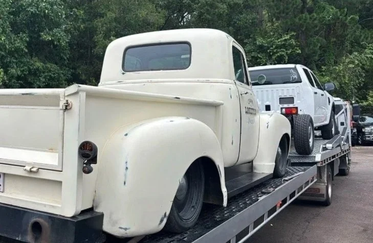 A white vintage pickup truck with a rounded cab is loaded on a flatbed tow truck, alongside a white classic Porsche 911.