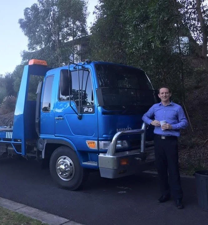 A man in a blue dress shirt and black pants standing next to a blue tow truck, holding a cup, outdoors with trees and a building in the background.