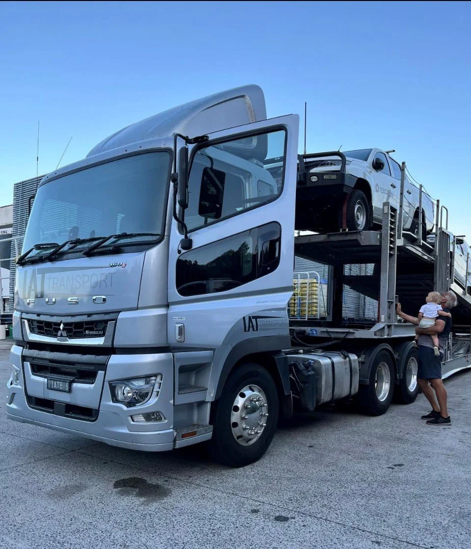 A car carrier truck carrying a black off-road vehicle, with a woman holding a child standing nearby.