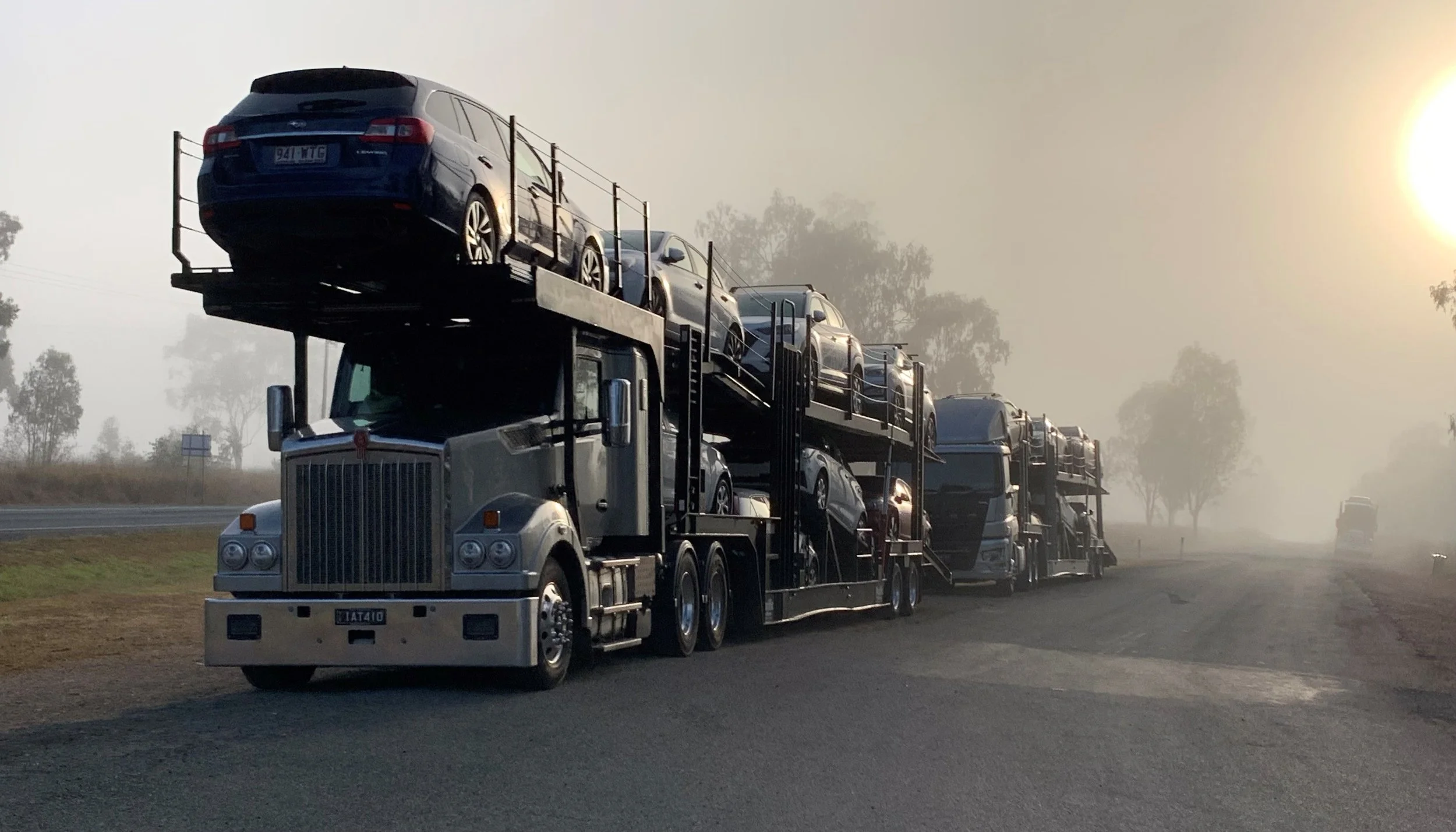 A car carrier truck with multiple cars loaded on two levels parked on the side of a road during dusk or dawn with mist and trees in the background.